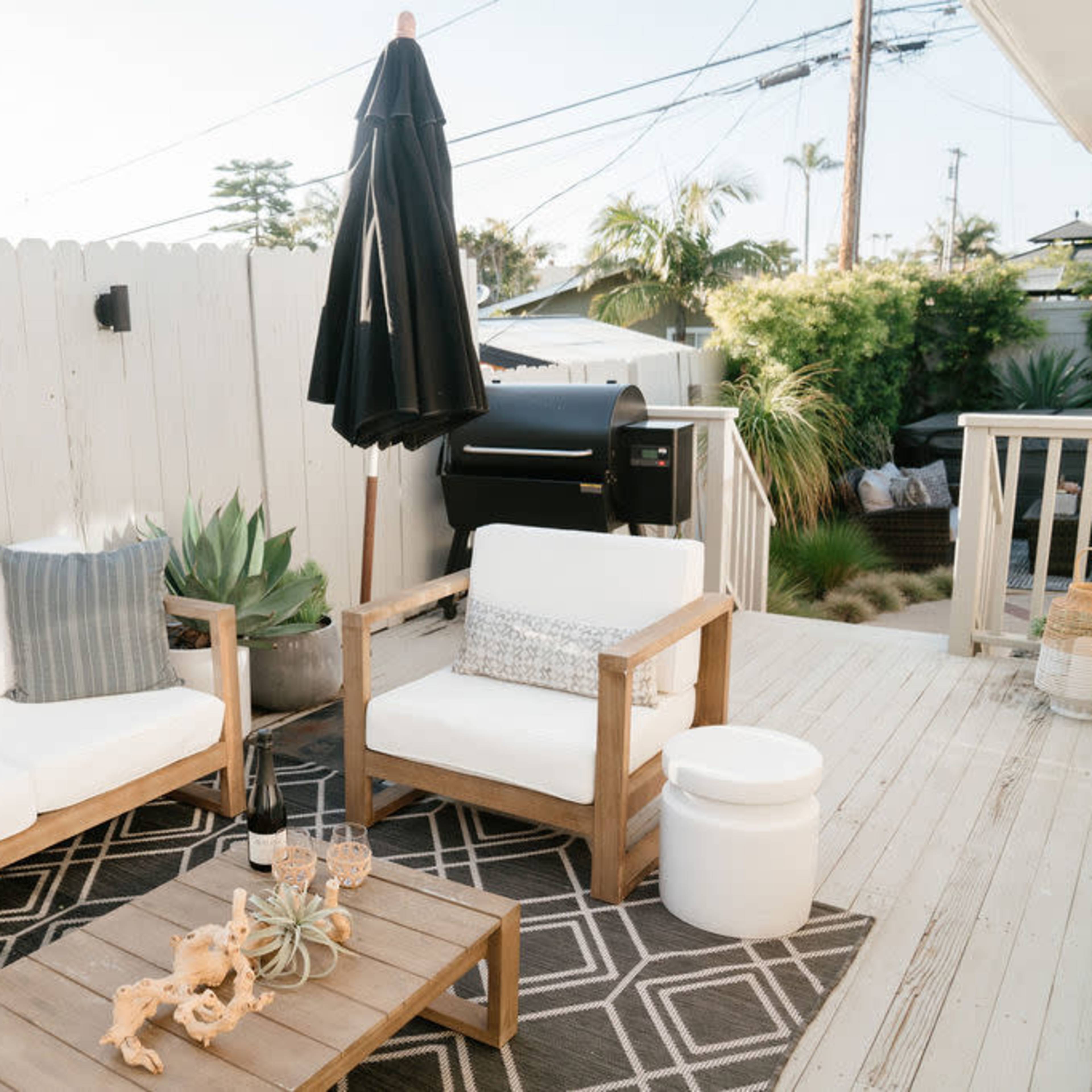 A patio area with a wooden table, white seating, a black umbrella, and greenery in the background.