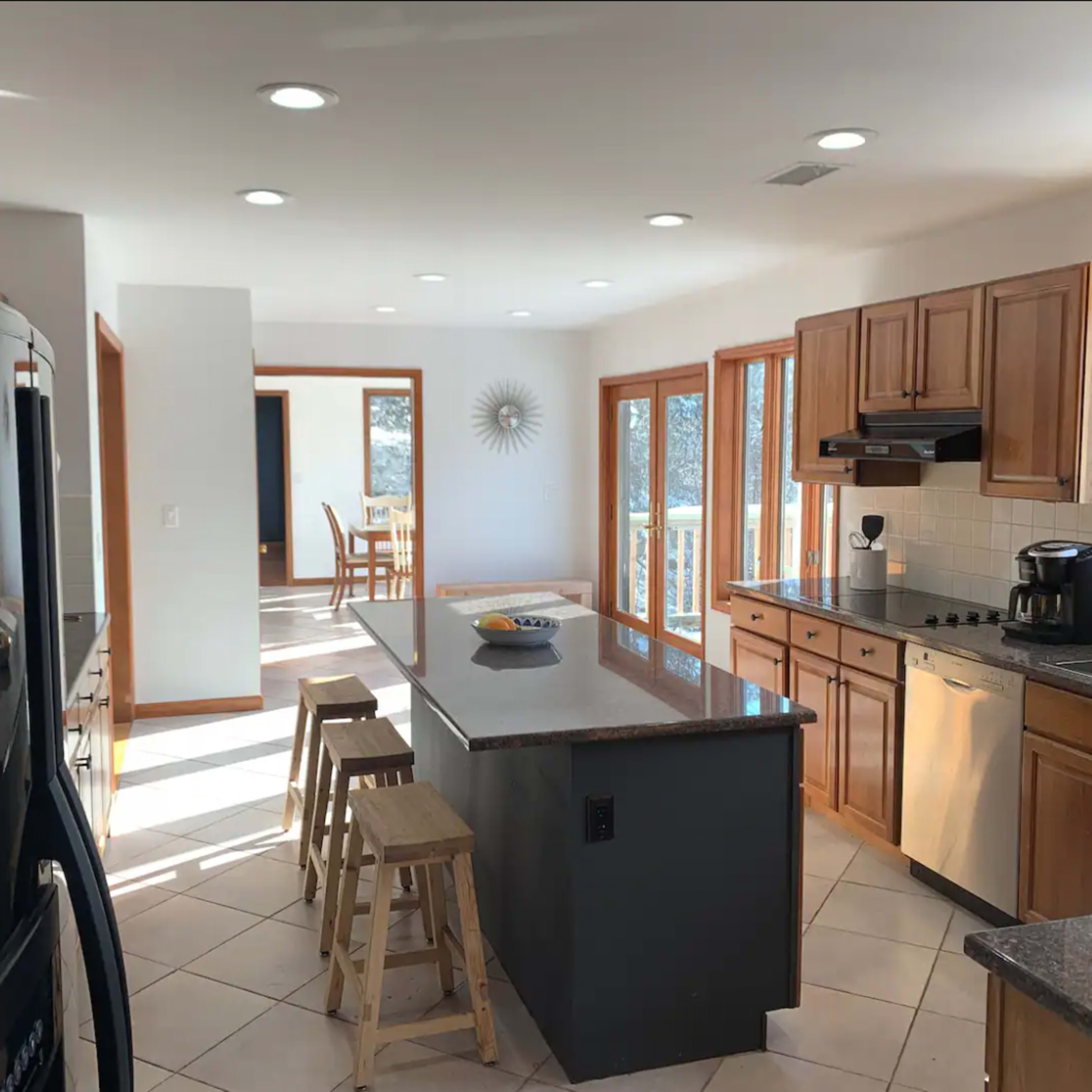 A modern kitchen features wooden cabinetry, a large island with stools, and windows that allow natural light to fill the space.
