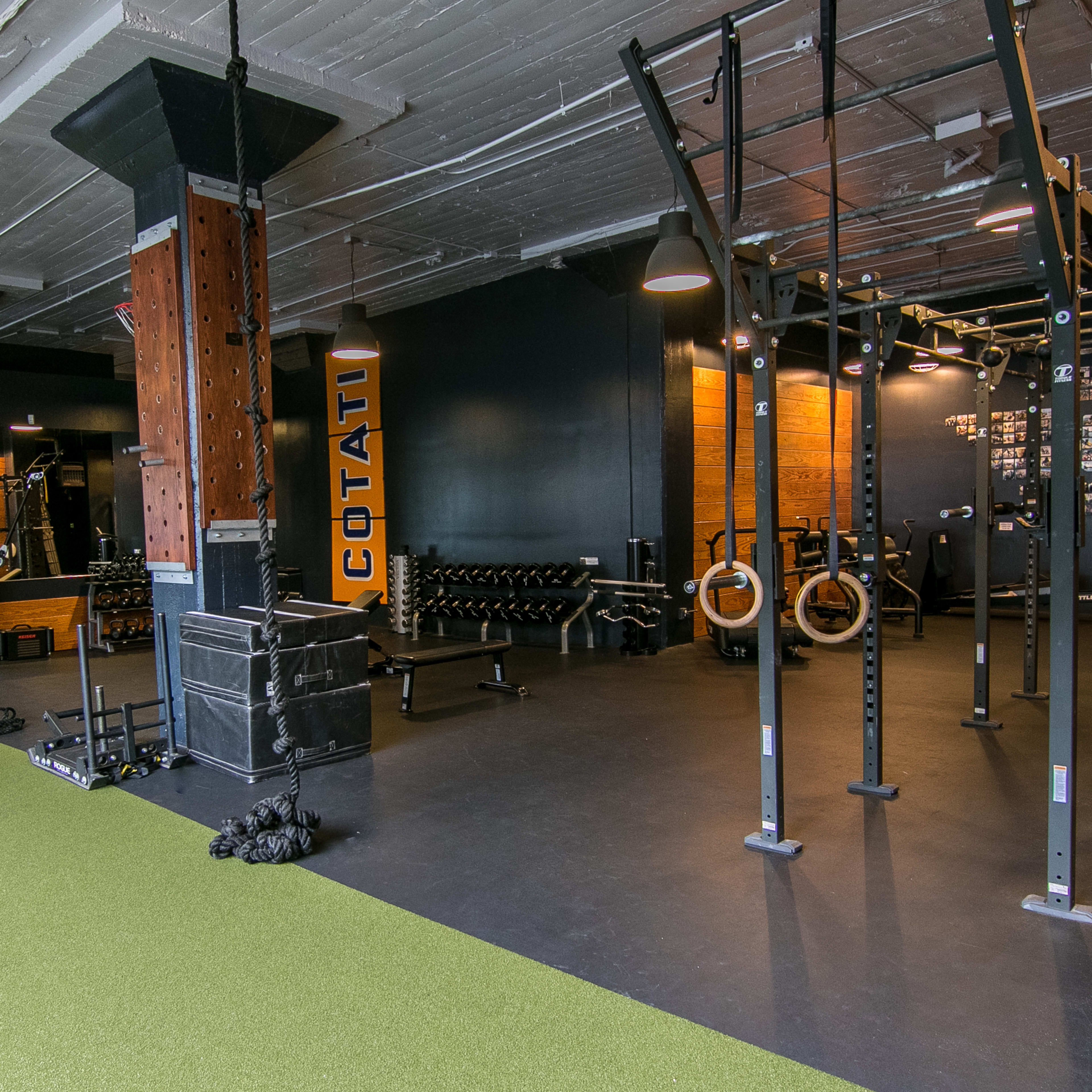 A well-equipped gym with various workout stations, including weightlifting areas, exercise machines, and resistance training equipment, all set against a backdrop of dark walls and green turf flooring.