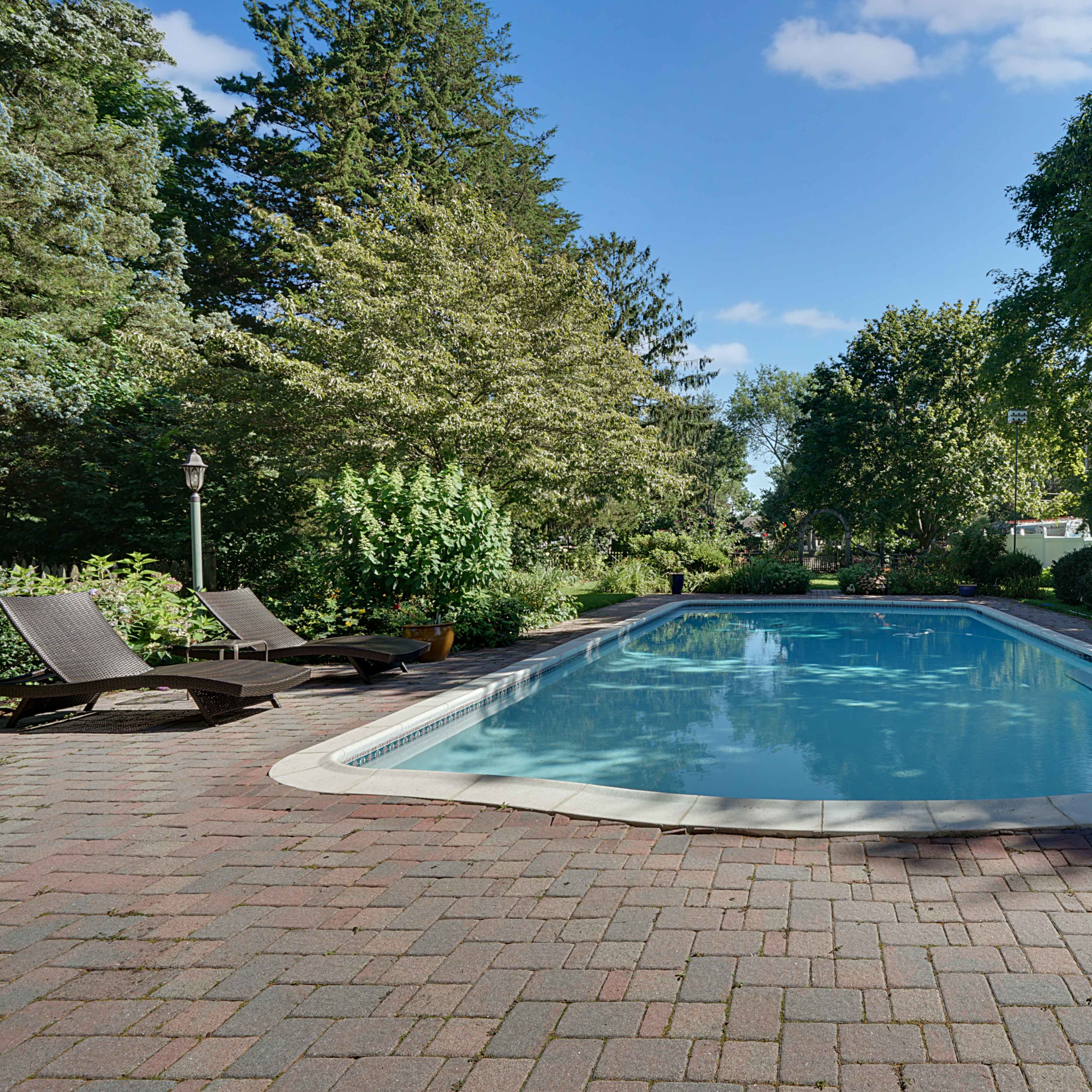 A rectangular swimming pool surrounded by a brick patio, lounge chairs, and lush greenery under a clear blue sky.