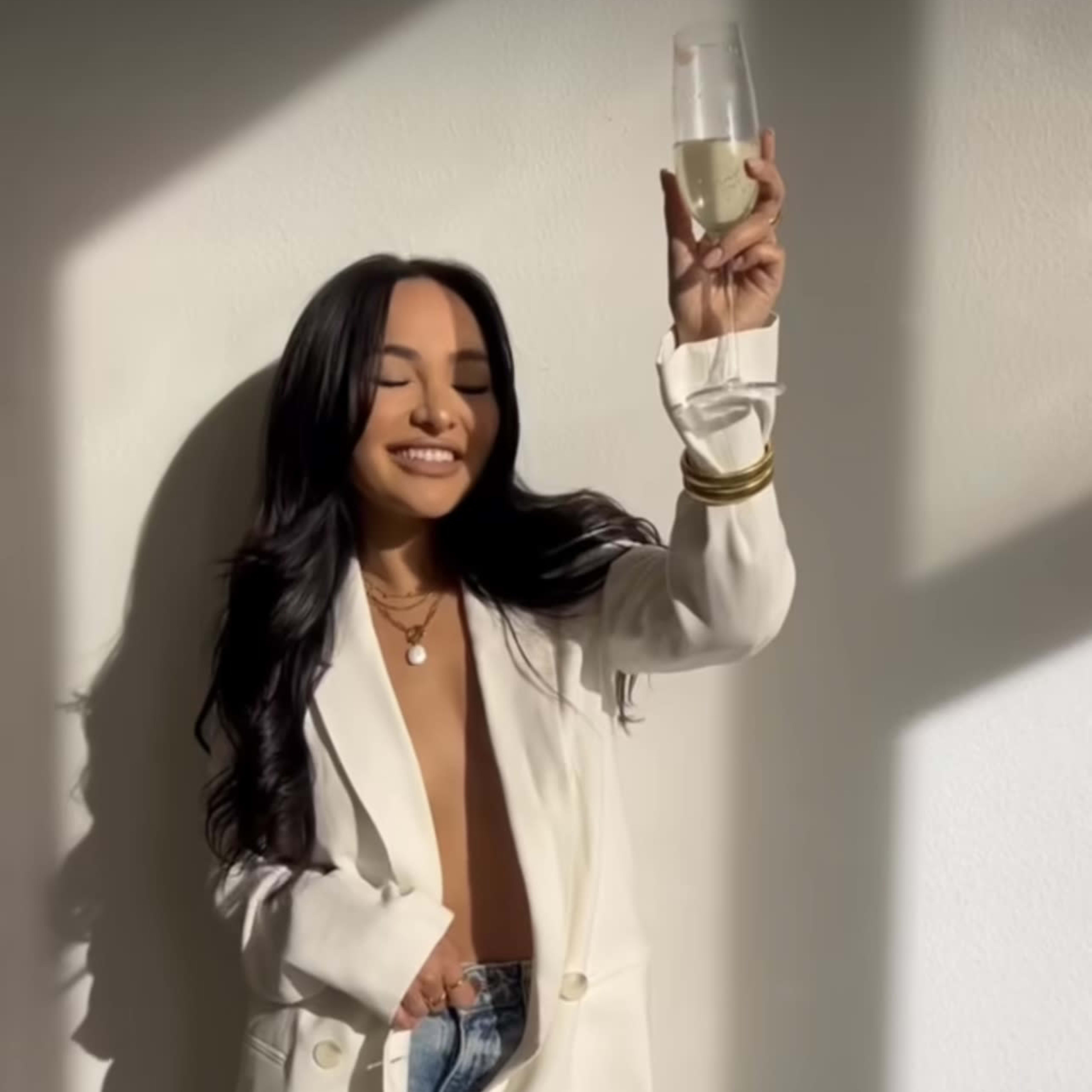 A woman in a white blazer holds a glass of champagne while leaning against a wall.