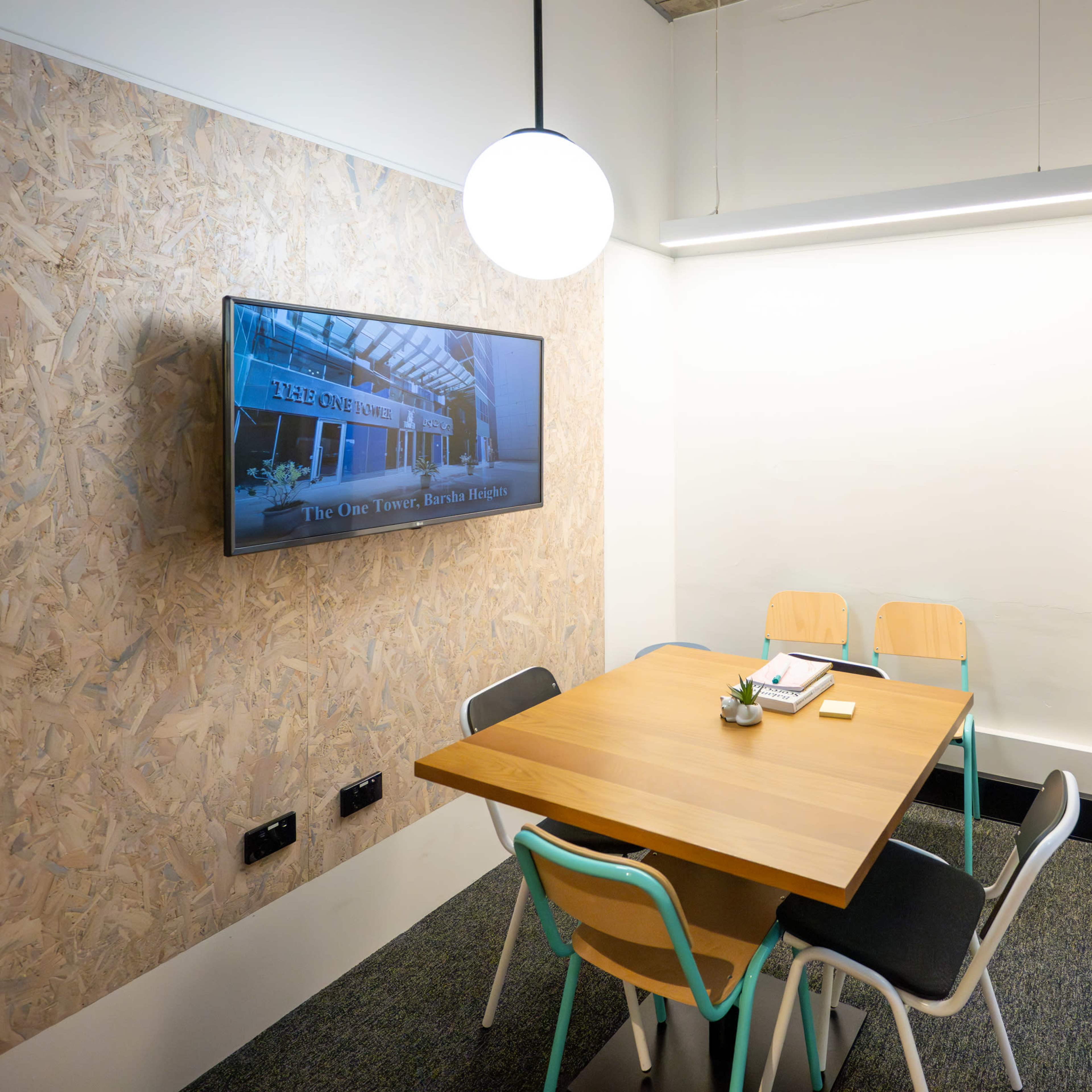 A small meeting room features a wooden table surrounded by four chairs, a wall-mounted television, and large windows that allow natural light.