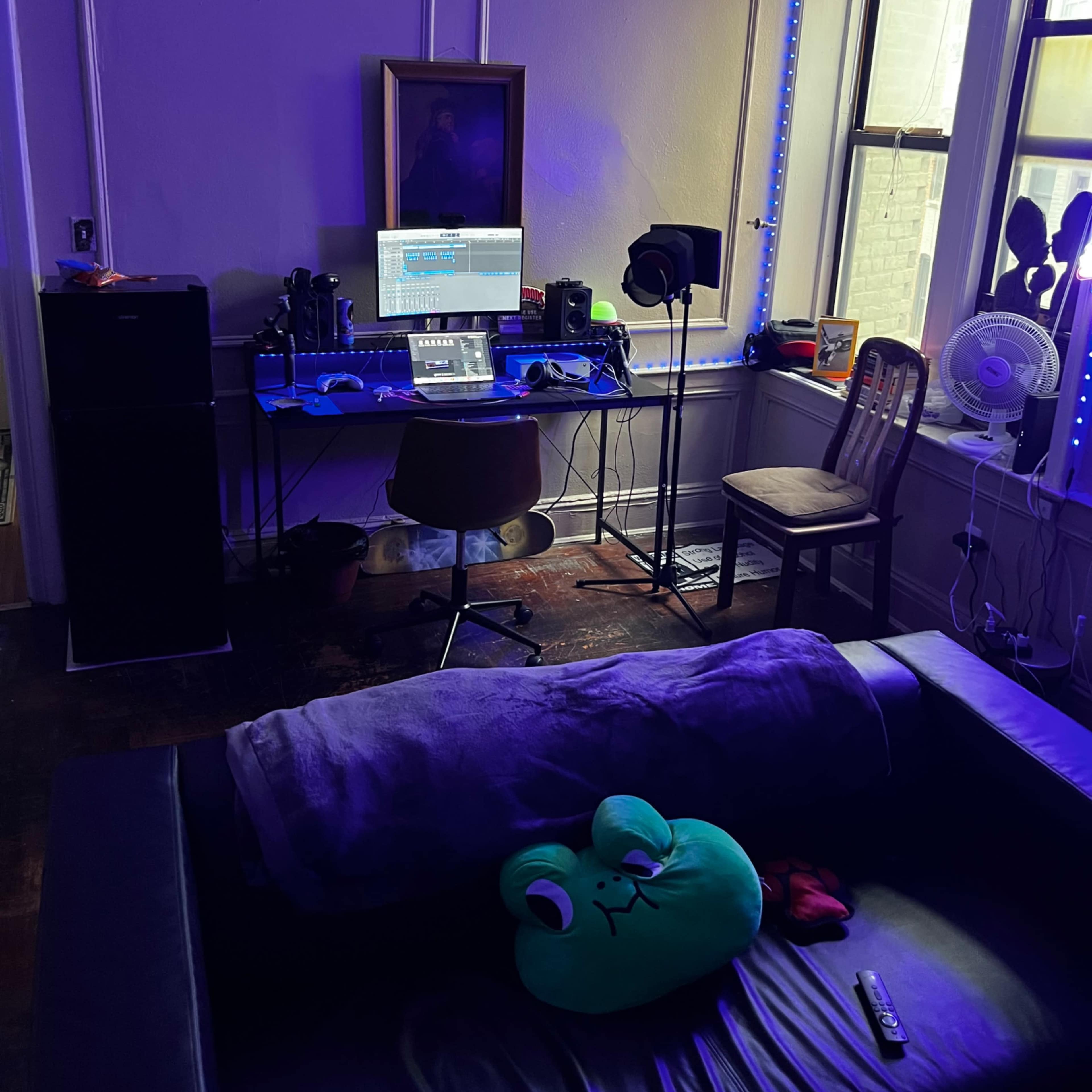 A room with a dark couch, a desk equipped with a computer and music production equipment, and ambient LED lights illuminating the walls.