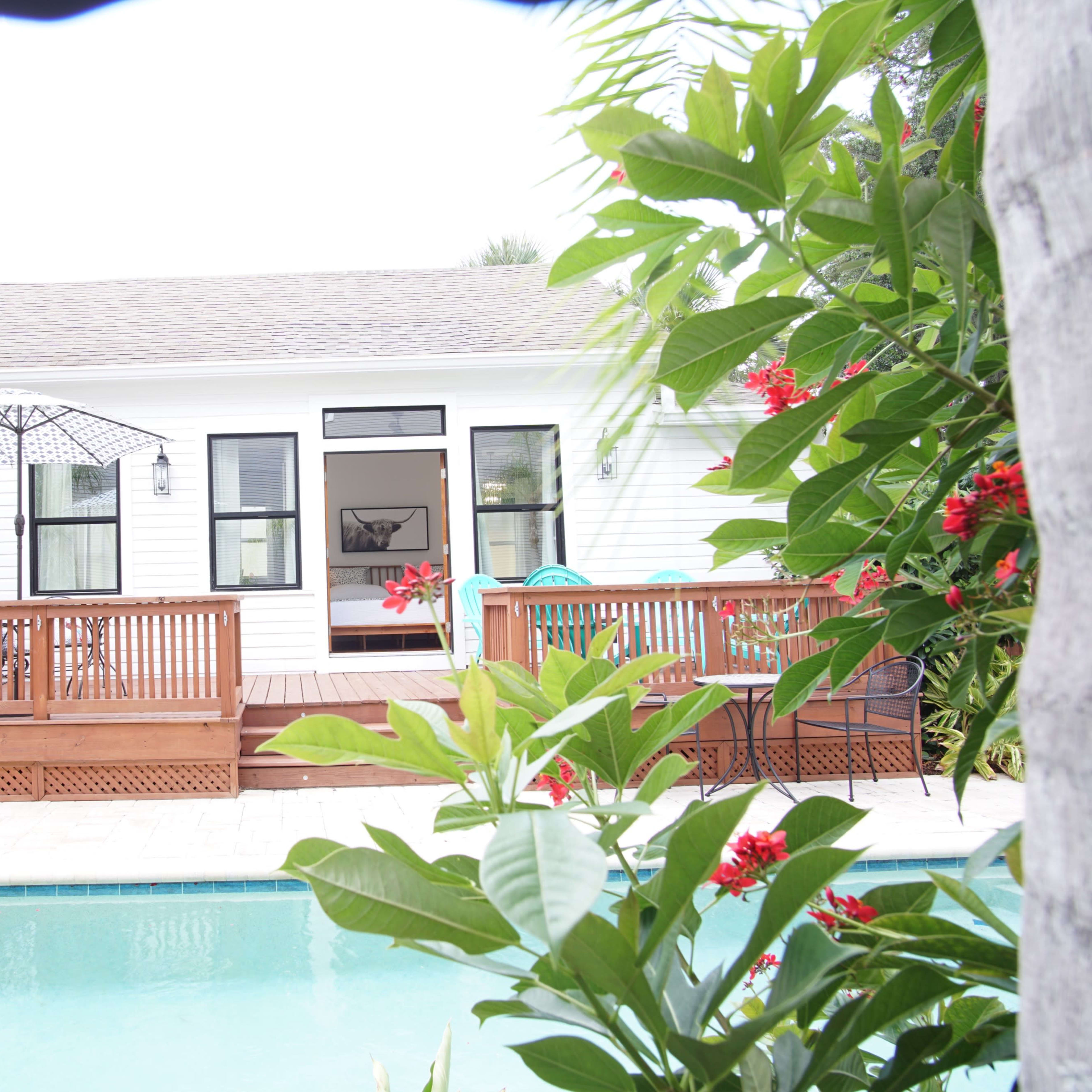A wooden deck leads to a white house with large windows, surrounded by lush greenery and a swimming pool in the foreground.