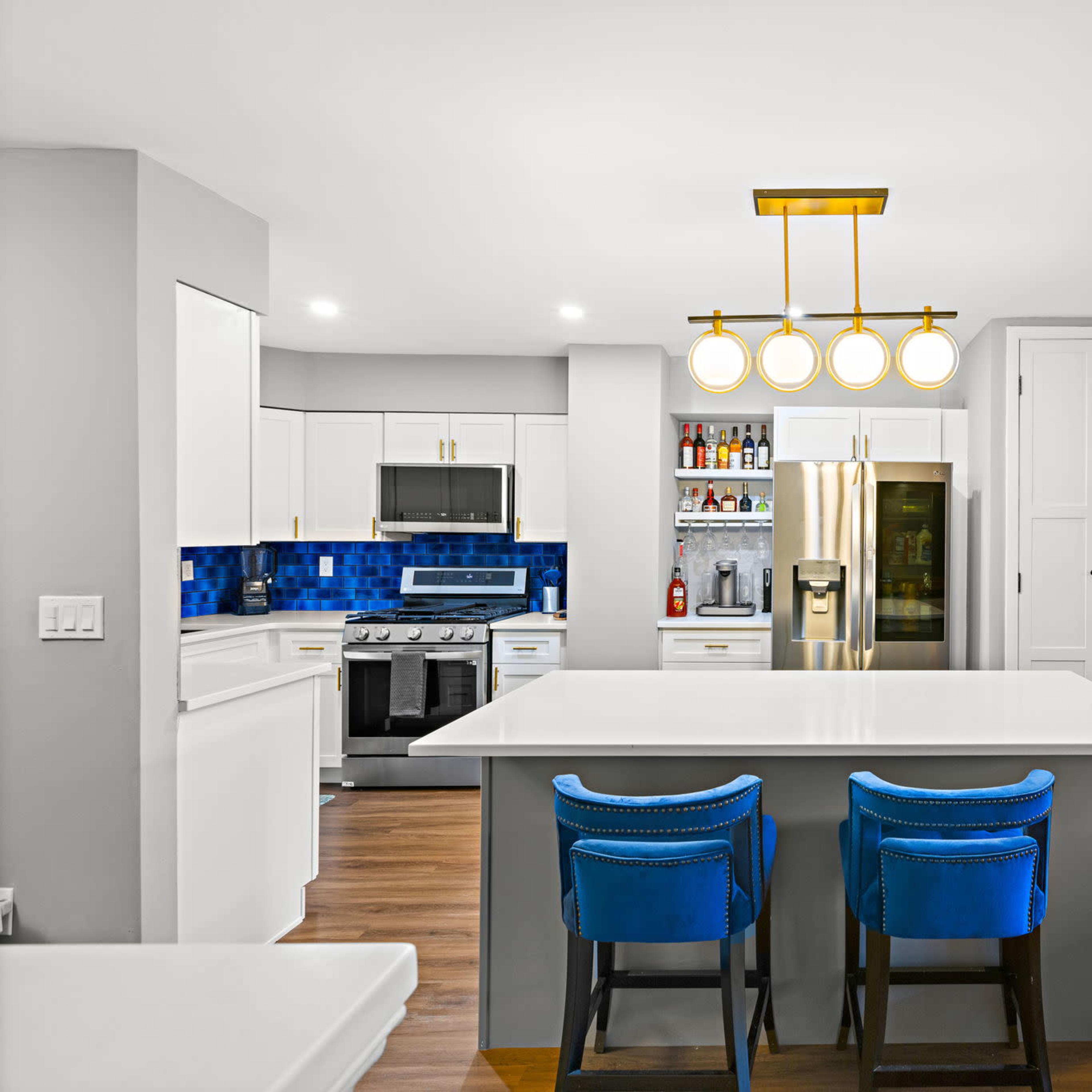 A modern kitchen features a large island with blue bar stools, stainless steel appliances, and blue tile backsplash.