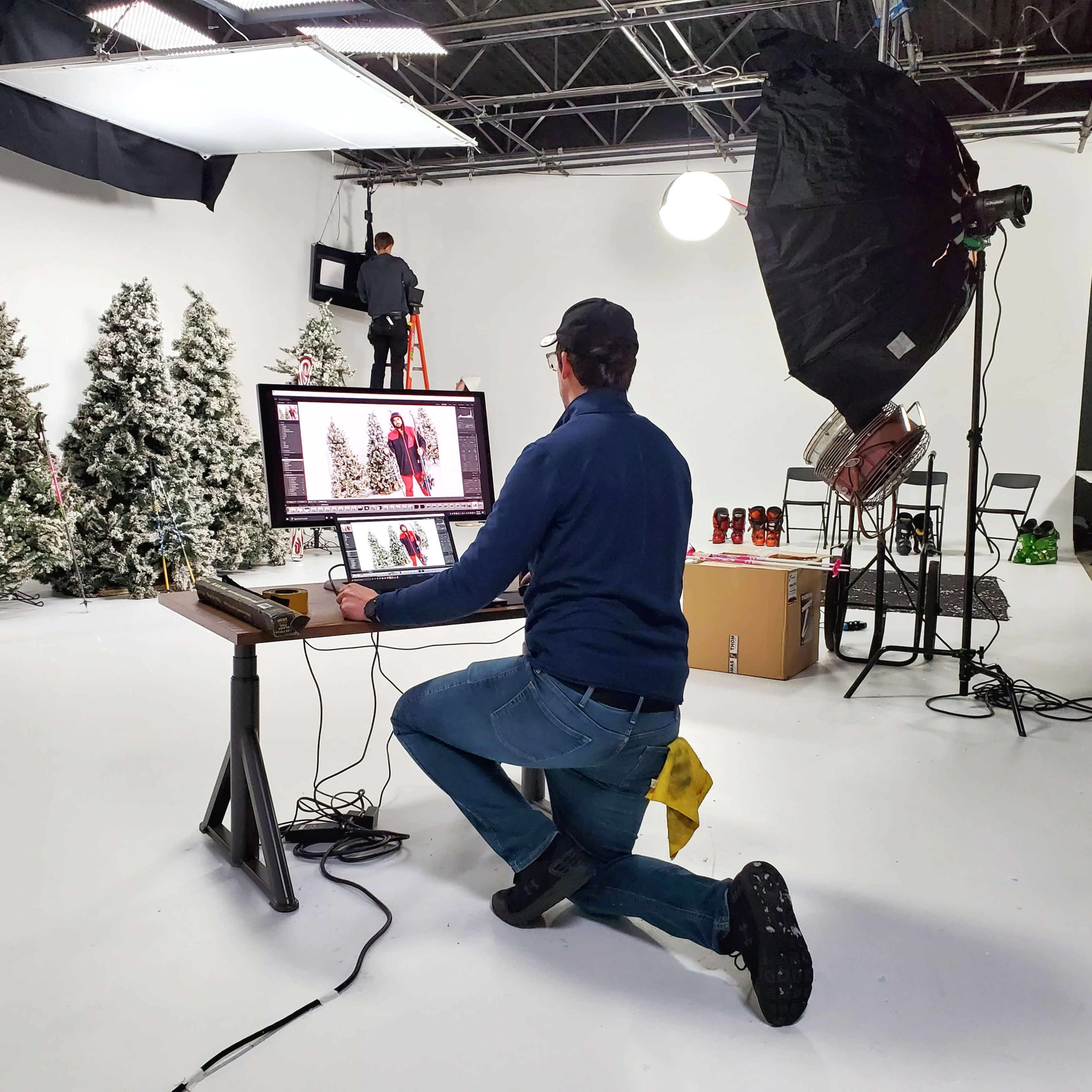 A photographer edits images on a computer while kneeling in a studio decorated with artificial Christmas trees and various equipment.