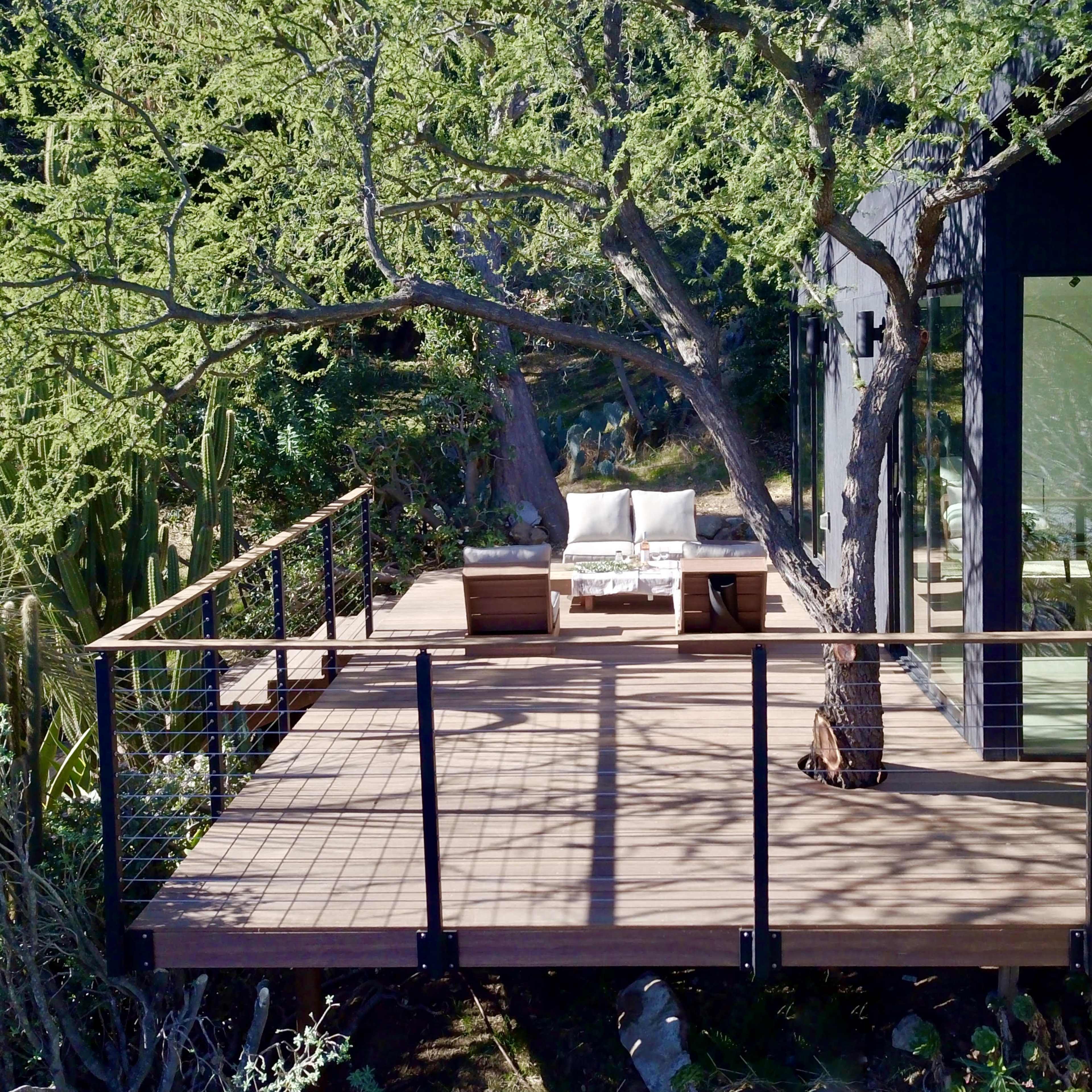A modern wooden deck features outdoor seating beside a house with large glass windows, surrounded by greenery and a prominent tree.