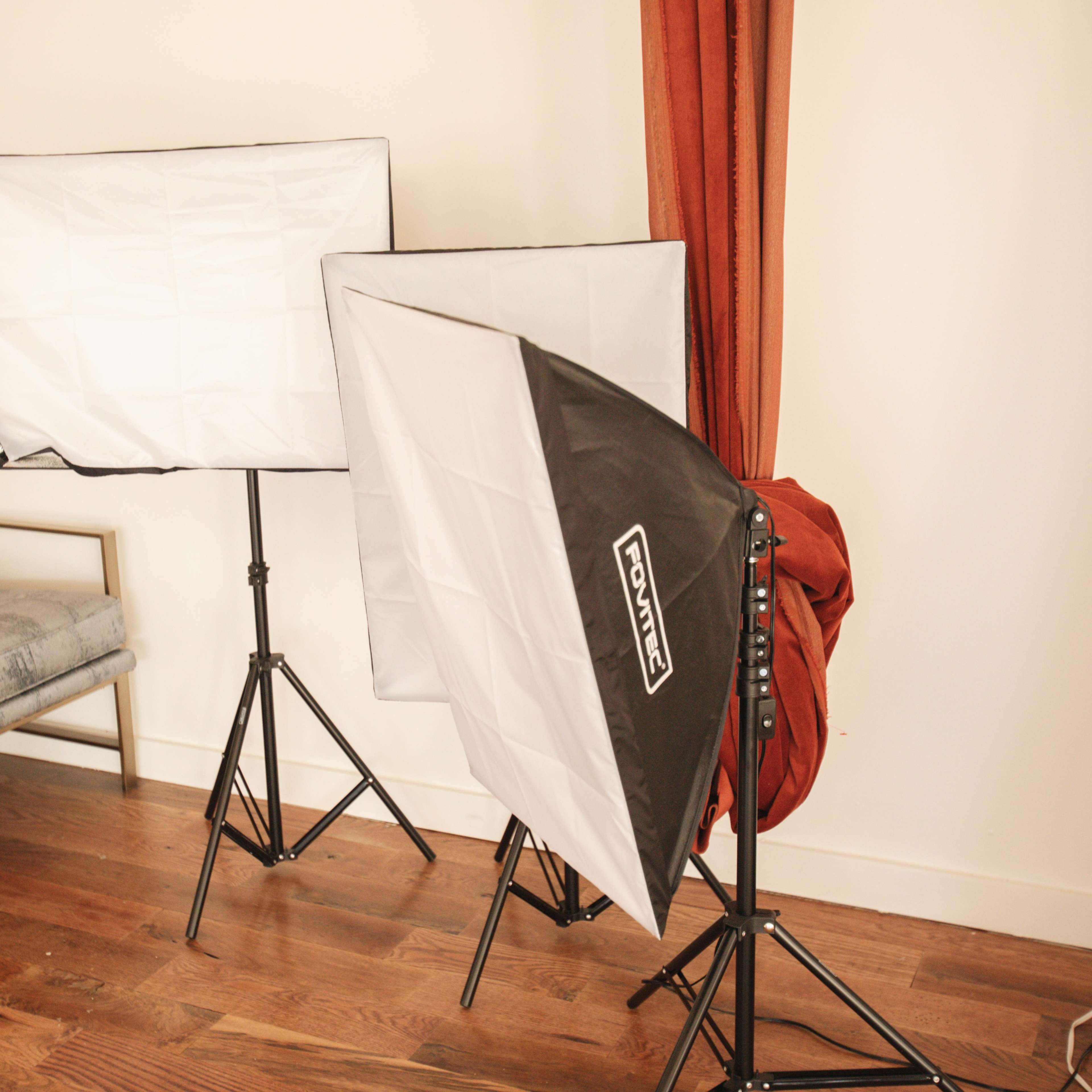 Three lighting softboxes set up on tripods in a room with wooden flooring and a fabric-covered chair.