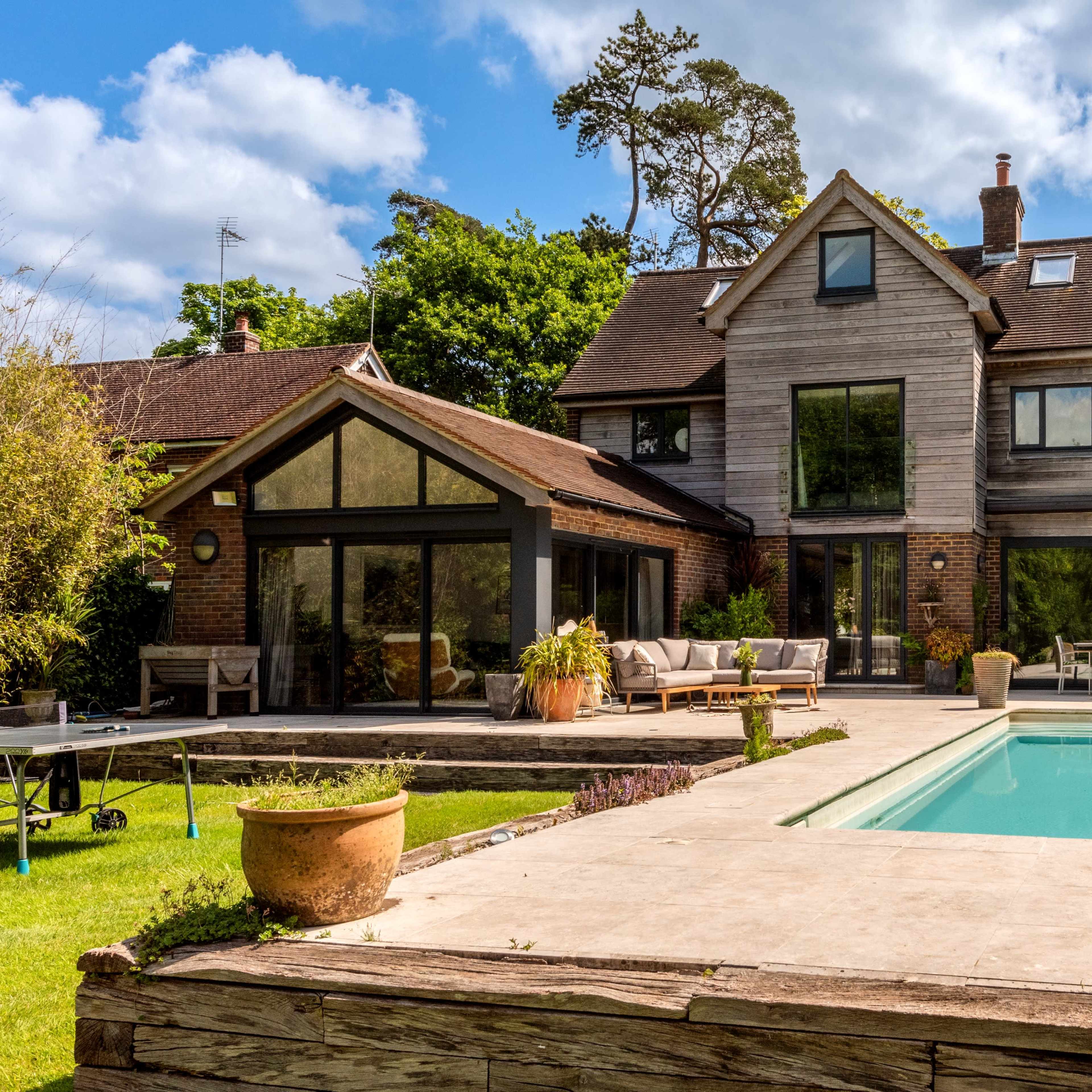 A modern two-story house with large windows, a patio area, and a swimming pool surrounded by greenery.