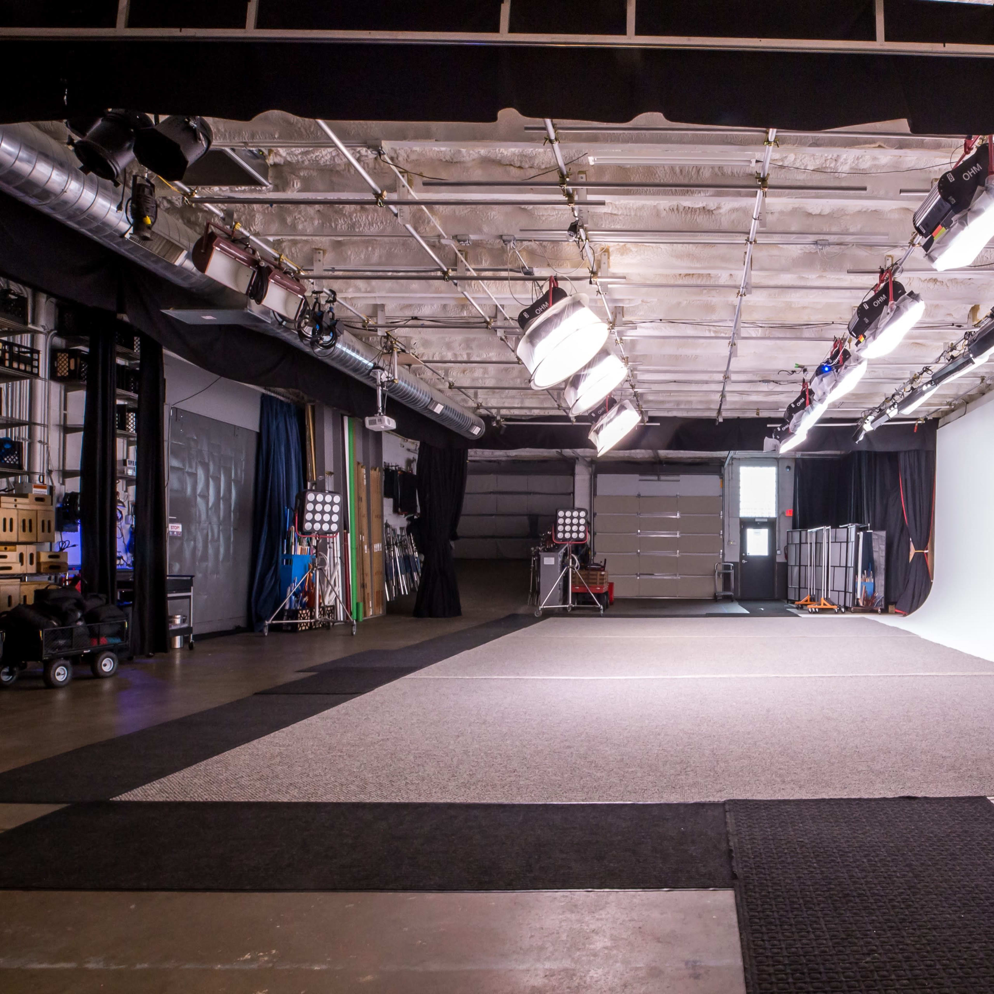 A spacious photography studio features a clean white backdrop, various lighting equipment, and multiple flooring surfaces.