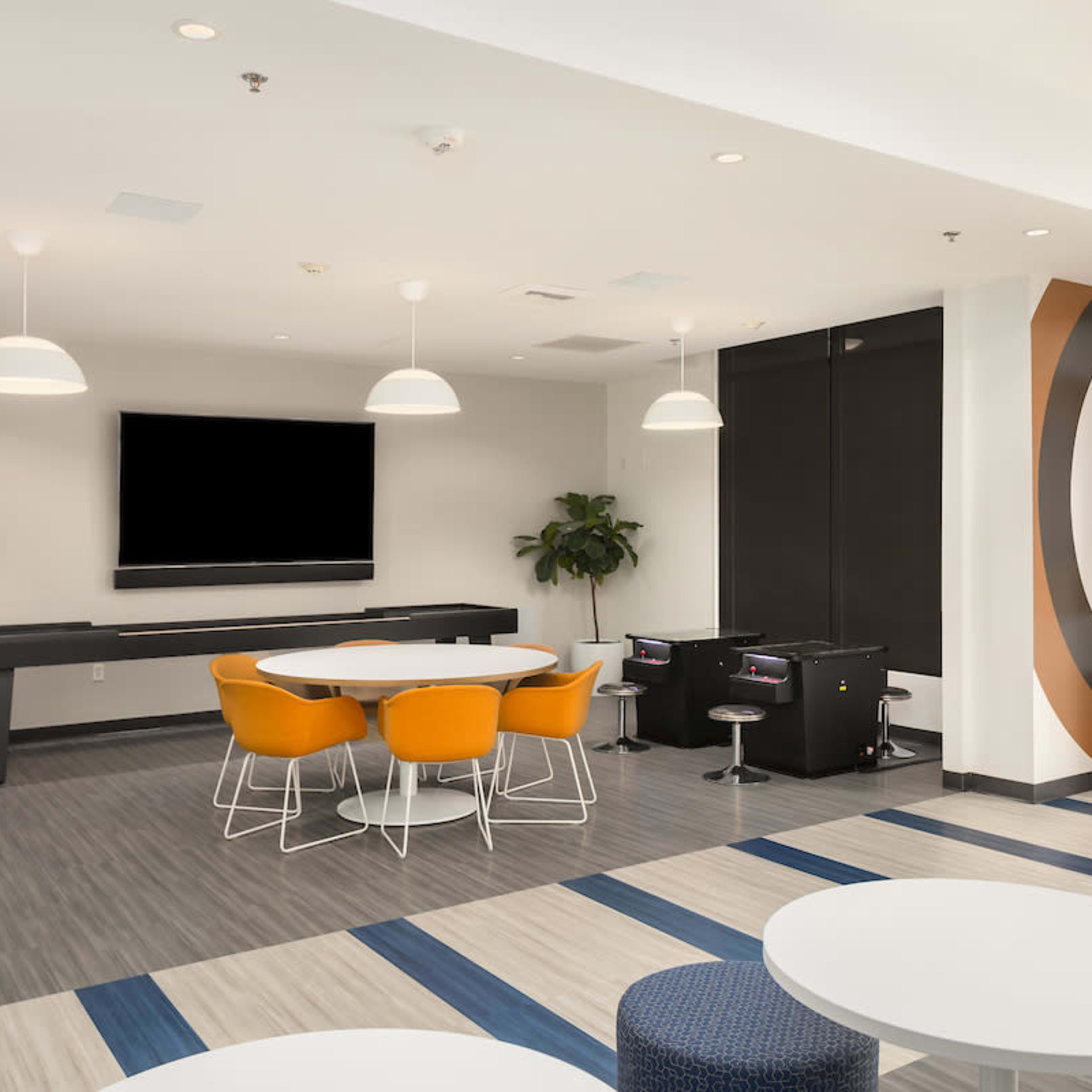A modern lounge area featuring circular tables, orange chairs, a wall-mounted television, and an abstract mural.