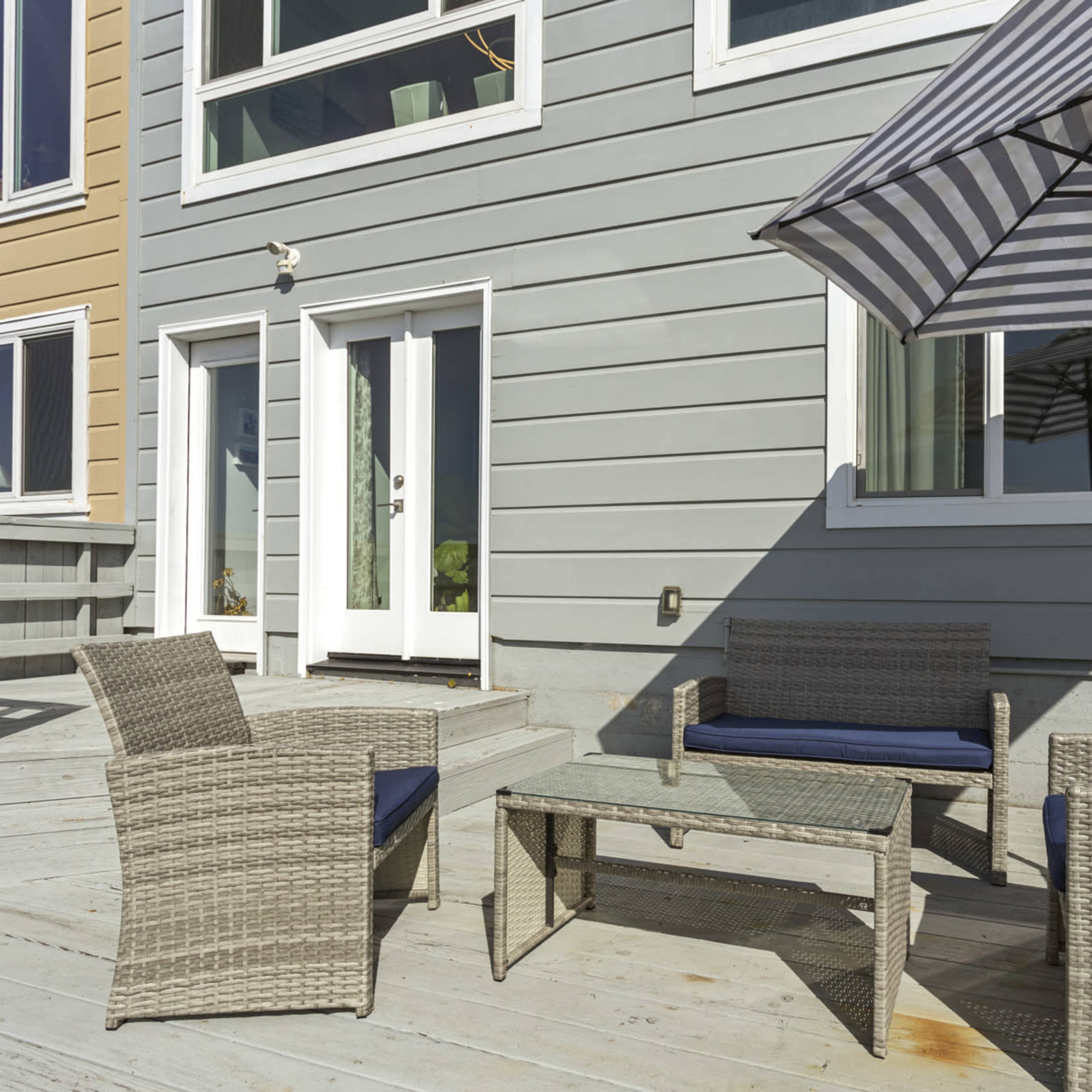 A seating area with wicker furniture and a striped umbrella is positioned on a wooden deck beside a gray house.