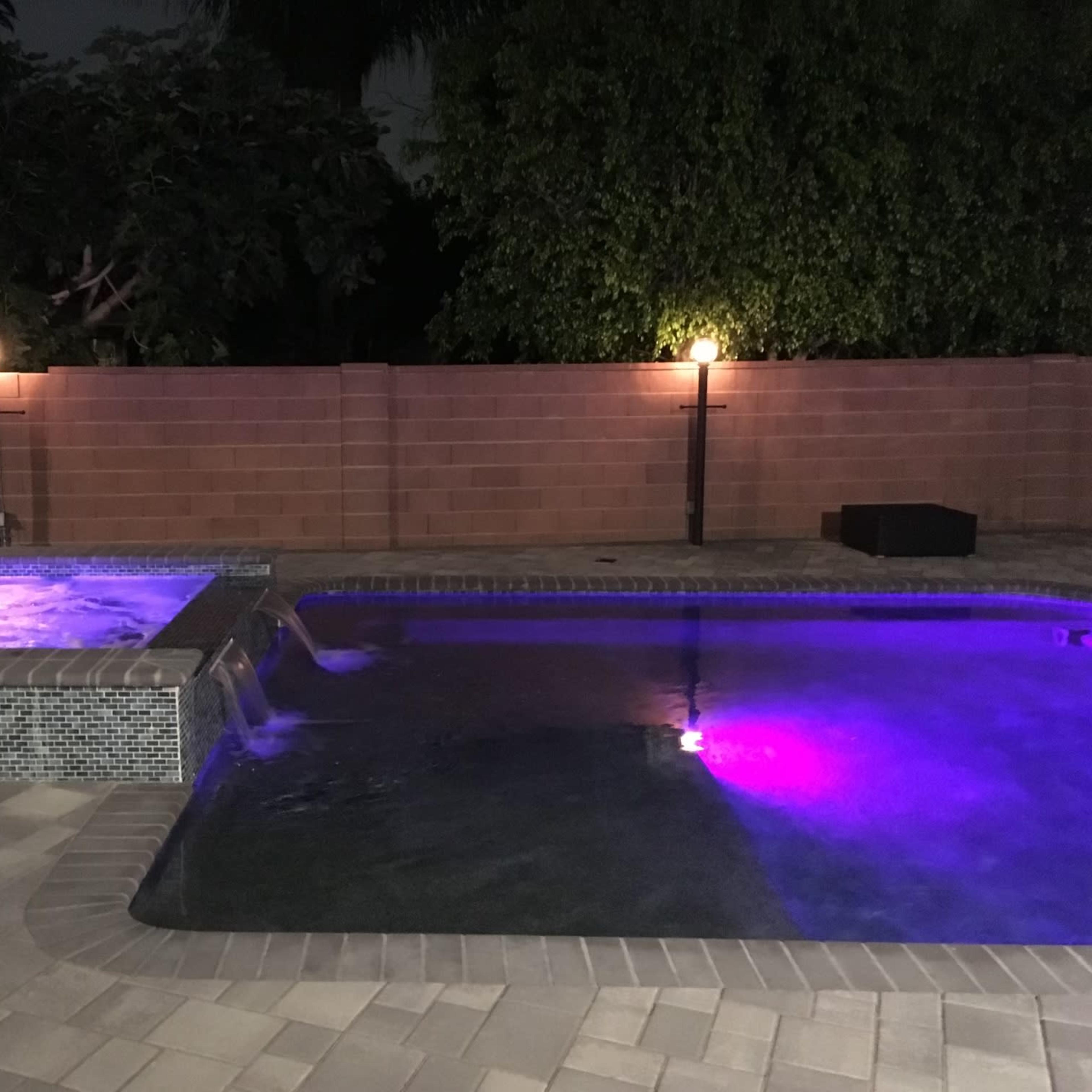 A swimming pool with a spa area is illuminated by purple lights against a brick wall backdrop.