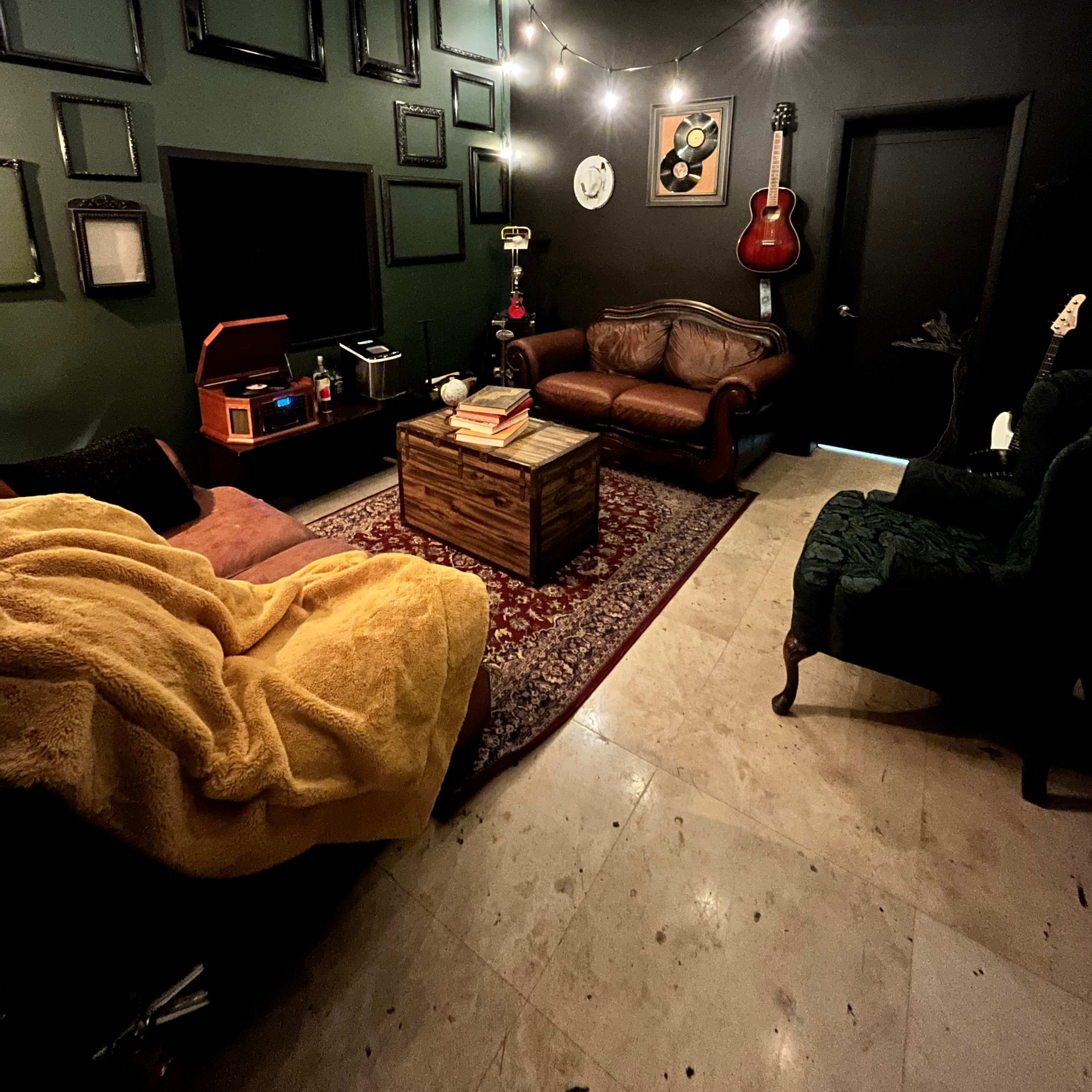 A cozy room features dark green walls adorned with empty picture frames, a sofa, a plush chair draped with a yellow blanket, and a wooden coffee table, all illuminated by string lights.