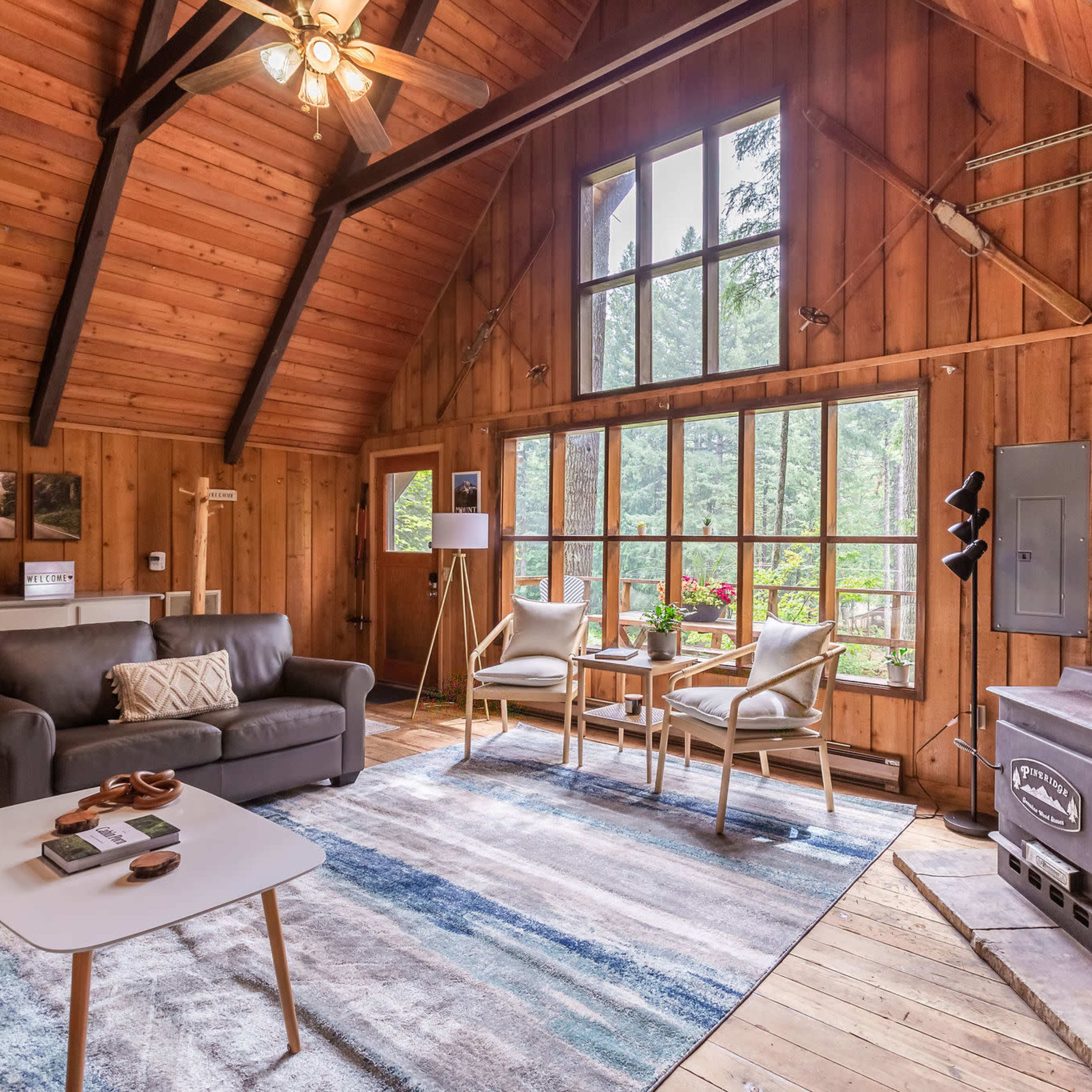 A cozy living room with wooden walls, large windows, a sofa, two chairs, and a wood-burning stove.