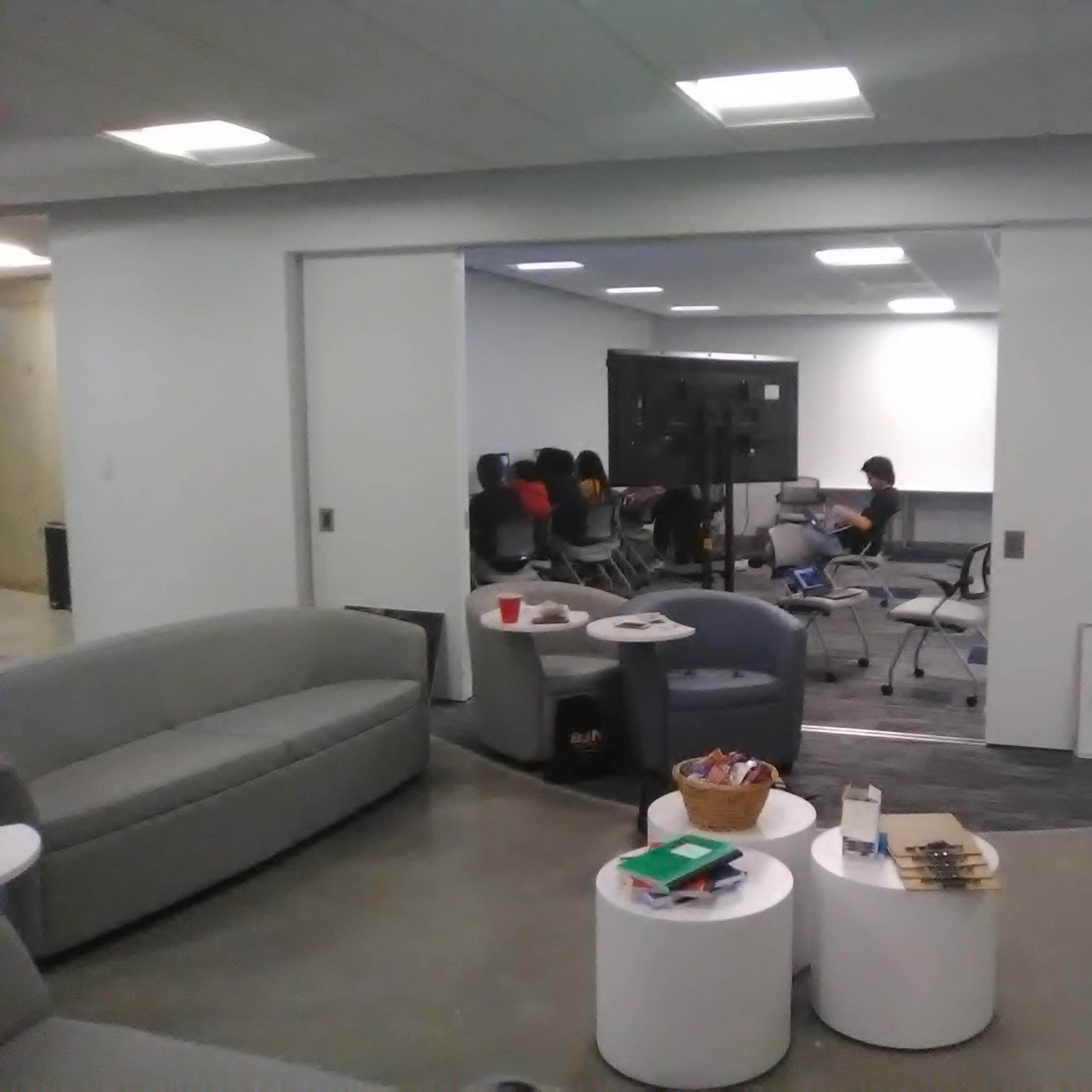 A group of people is seated in a conference room while a seating area with couches and tables is visible in the foreground.