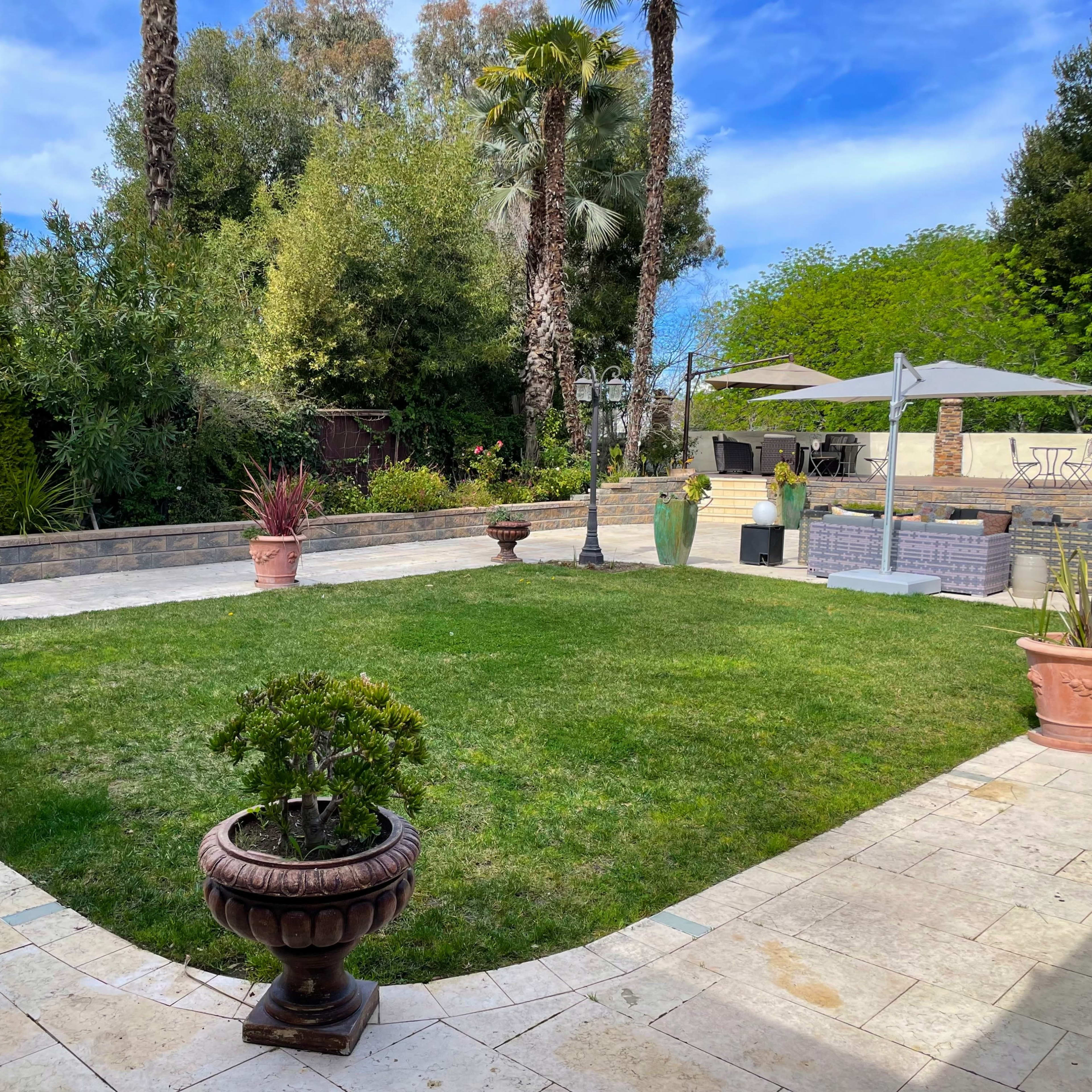 A landscaped garden with a grassy area surrounded by stone paths, decorative plants in pots, and outdoor furniture under a patio umbrella.