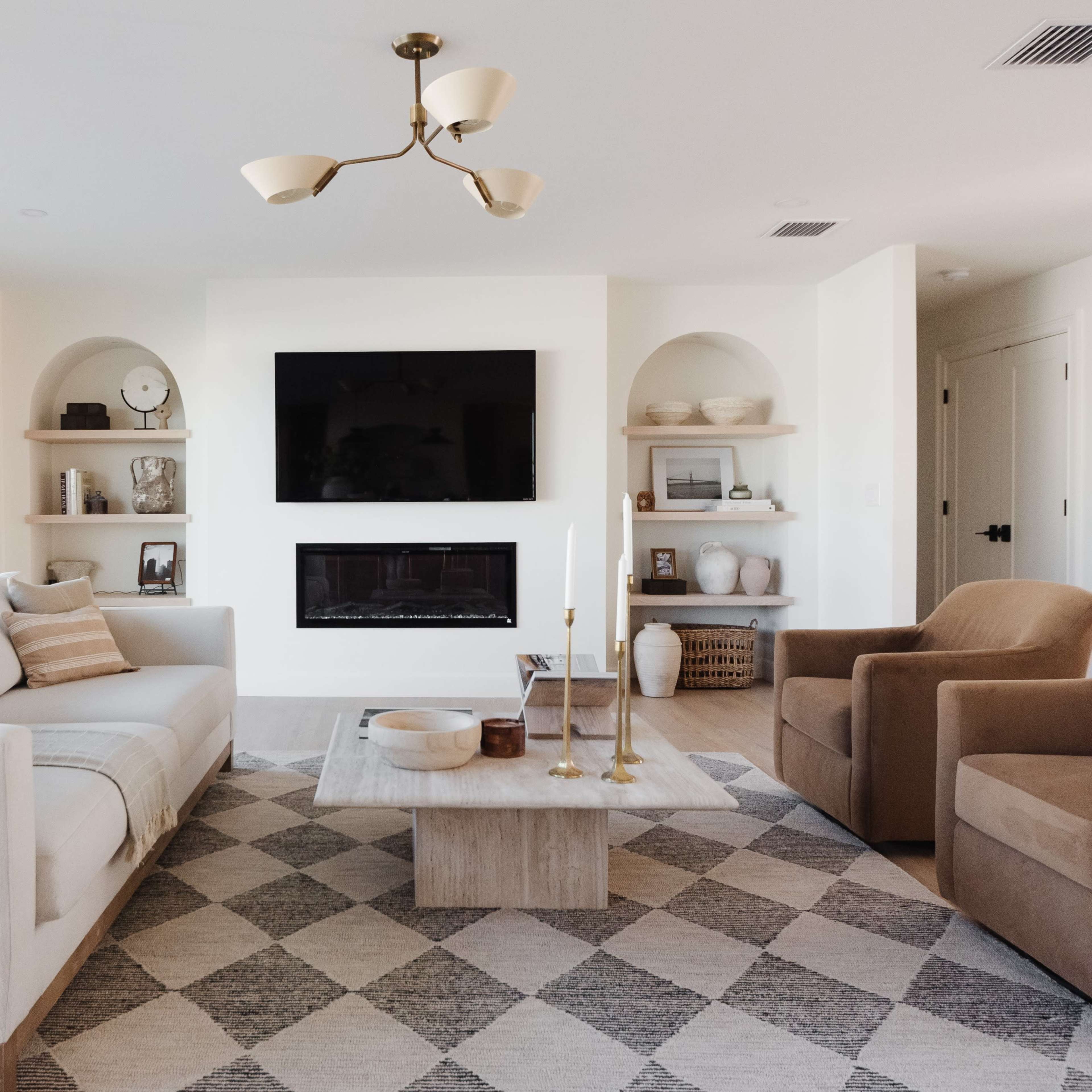 The living room features a sofa, two armchairs, a coffee table, and shelves with decorative items, all arranged around a modern fireplace.