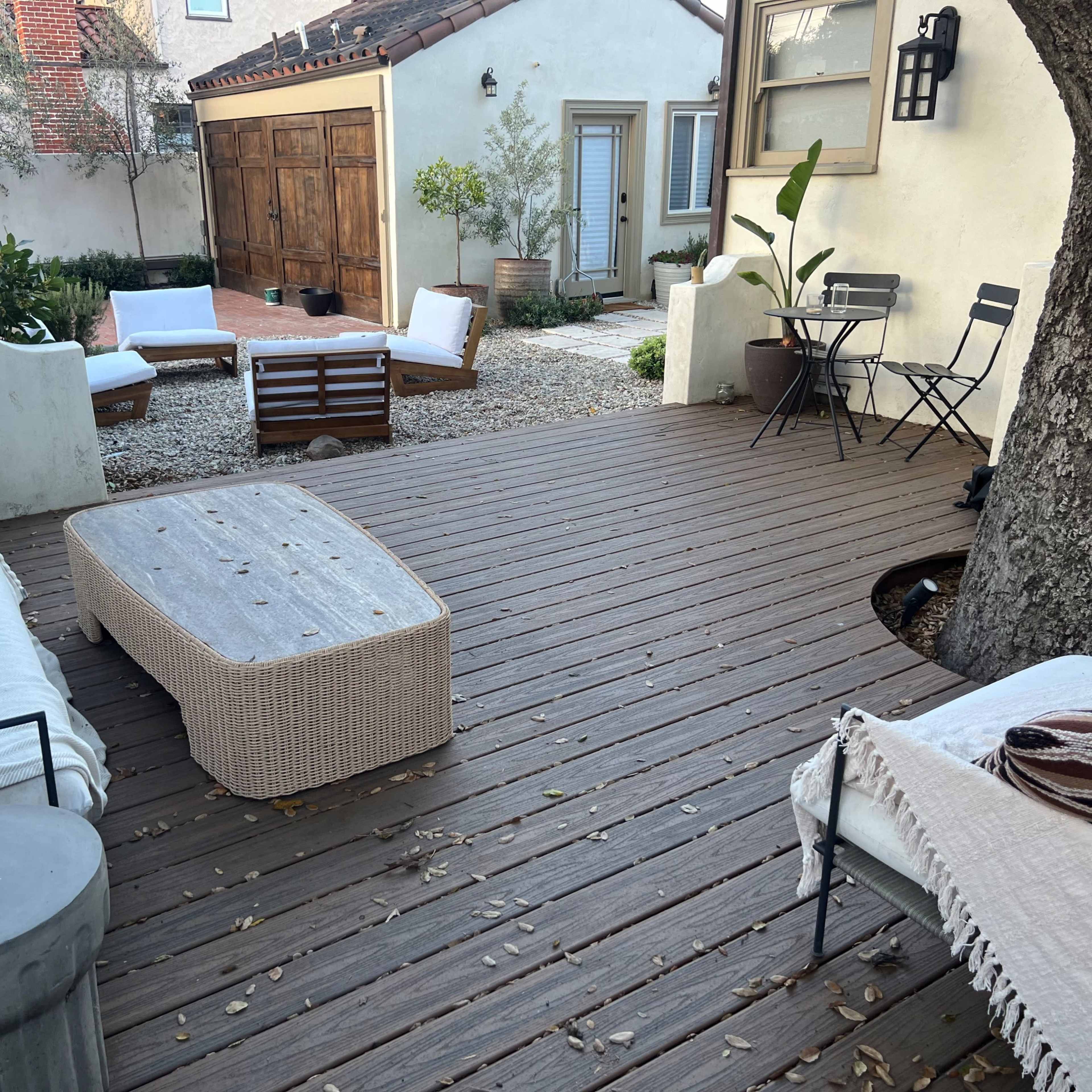 A patio area with wooden decking, featuring a coffee table, two lounge chairs, and a small bistro set, surrounded by potted plants and a gravel garden.