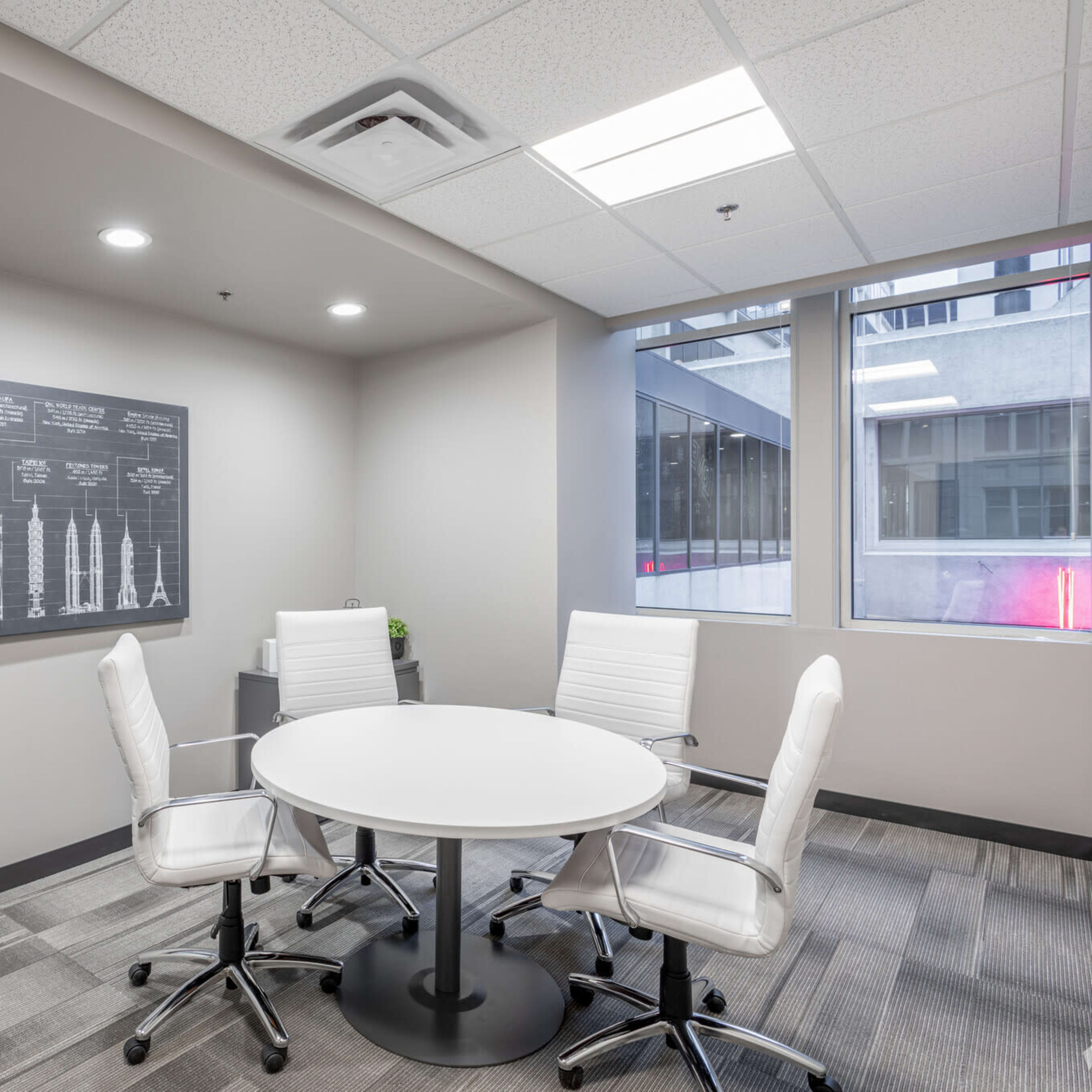A small meeting room with a round table and four white chairs, accompanied by a black wall chart and large windows.