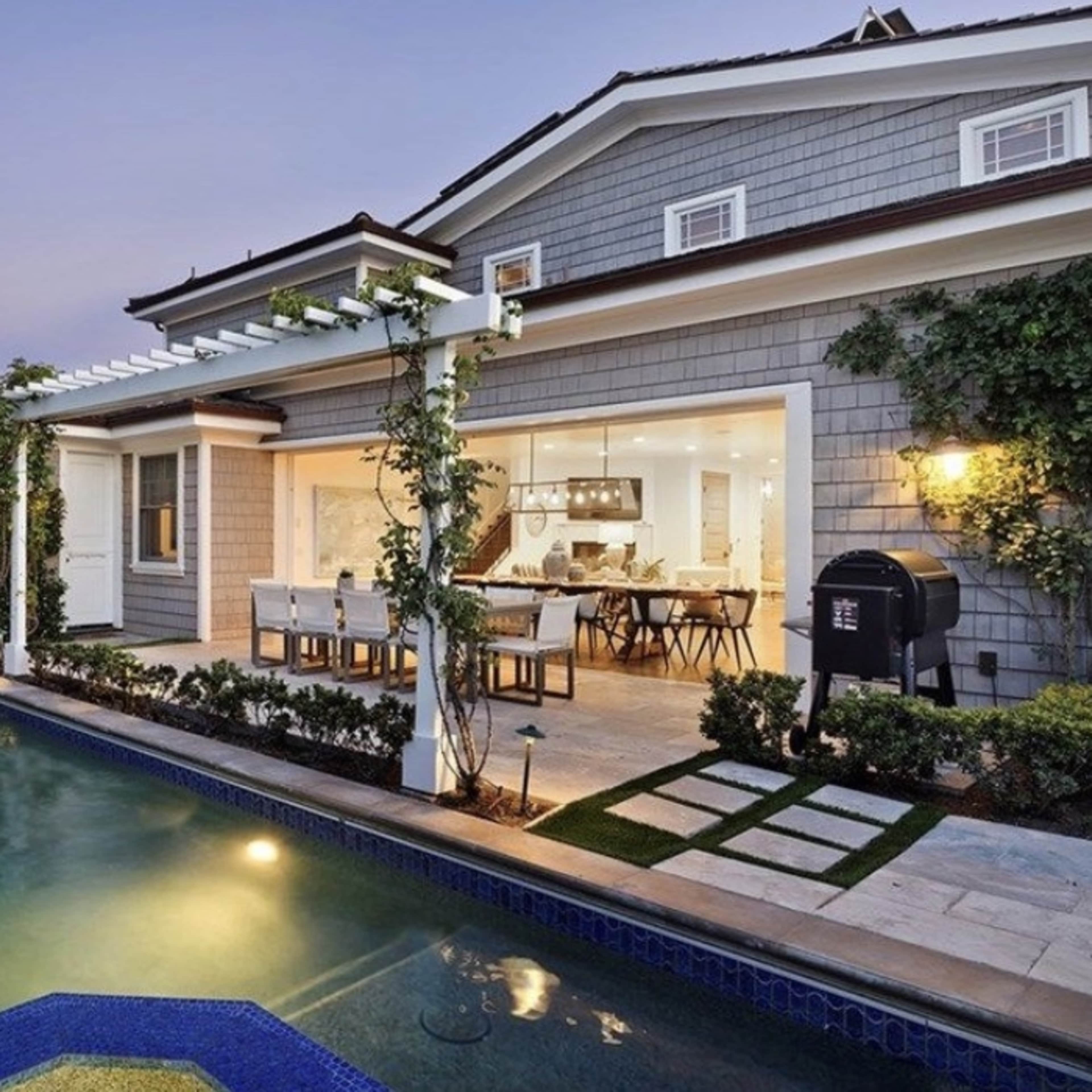 A modern house with a pool and landscaped garden features an outdoor dining area and large windows that provide a view into the interior.