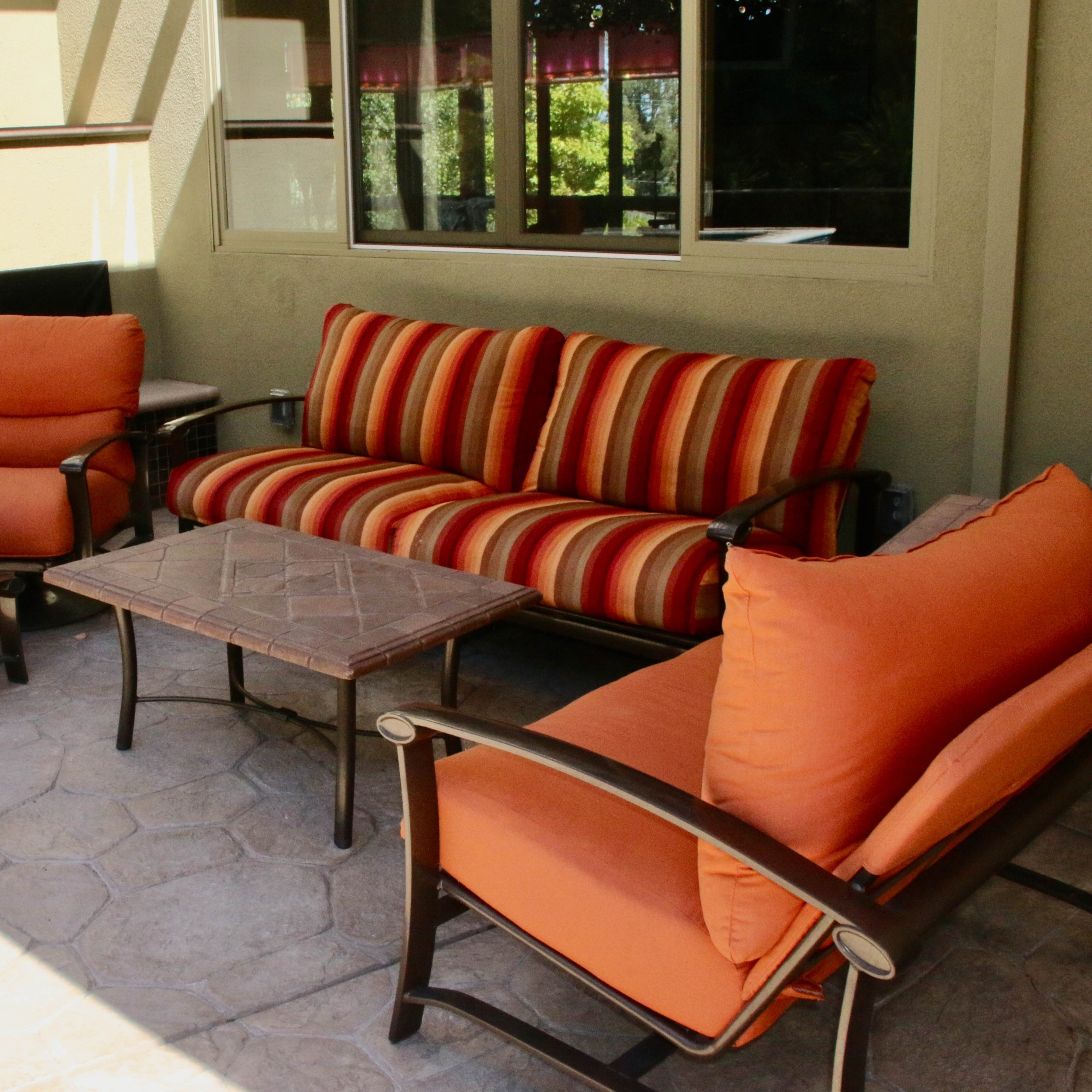 A patio seating area with a striped sofa, two orange chairs, and a coffee table on a textured stone floor.