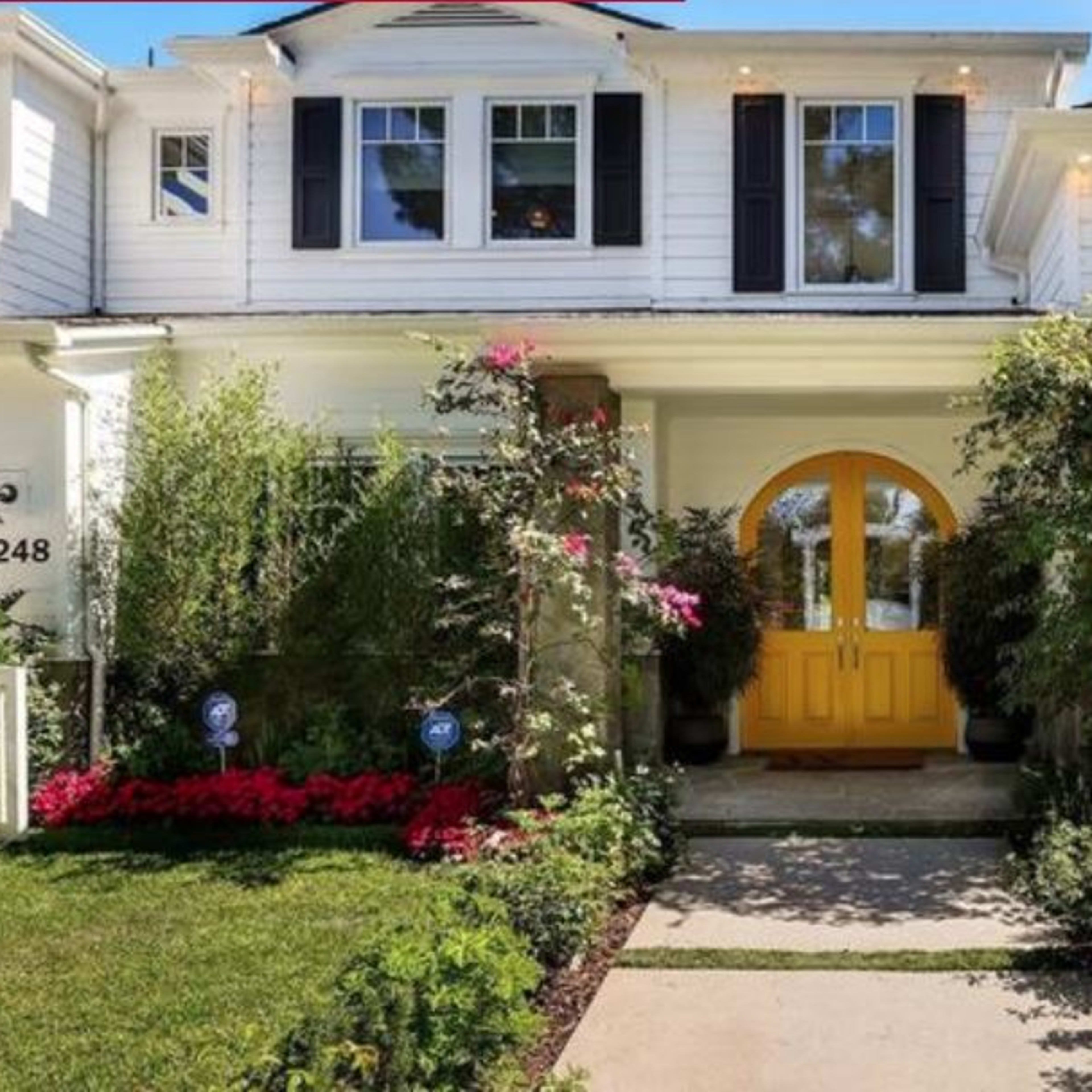 A white two-story house features a bright yellow front door, surrounded by landscaped gardens and a stone pathway.