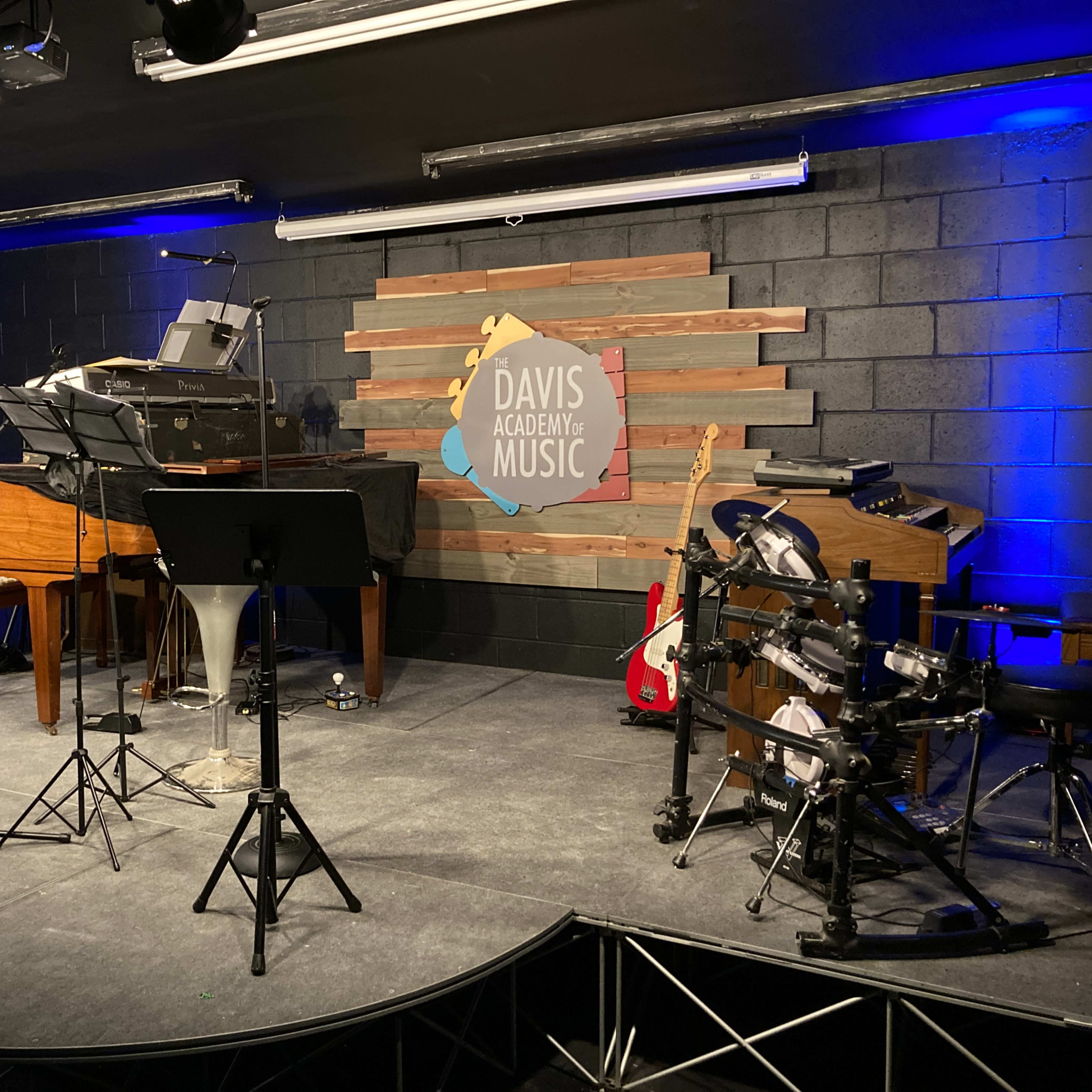 A music performance stage featuring a piano, a drum set, and various music stands, with a logo for Davis Academy of Music displayed on the back wall.
