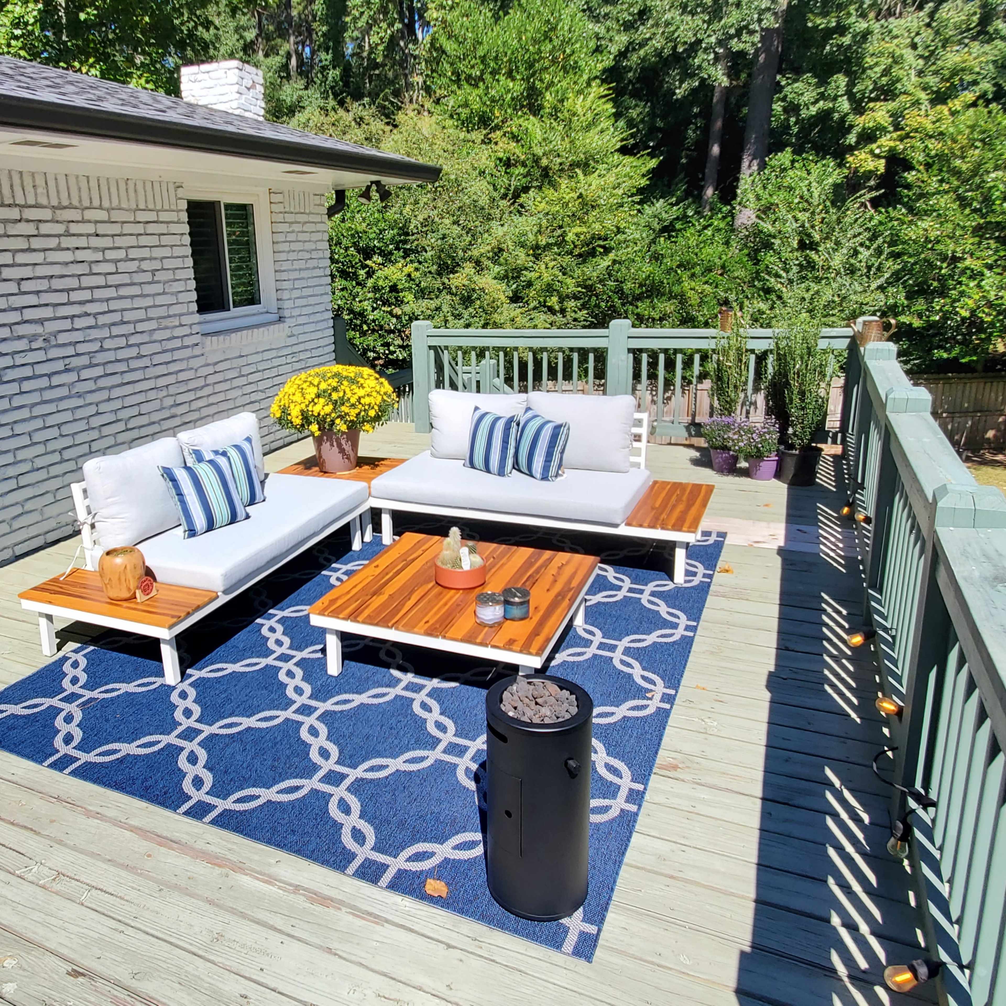 A patio area features two white sofas with blue and white striped cushions, a wooden coffee table, and a black fire pit, all set on a blue patterned rug surrounded by greenery.