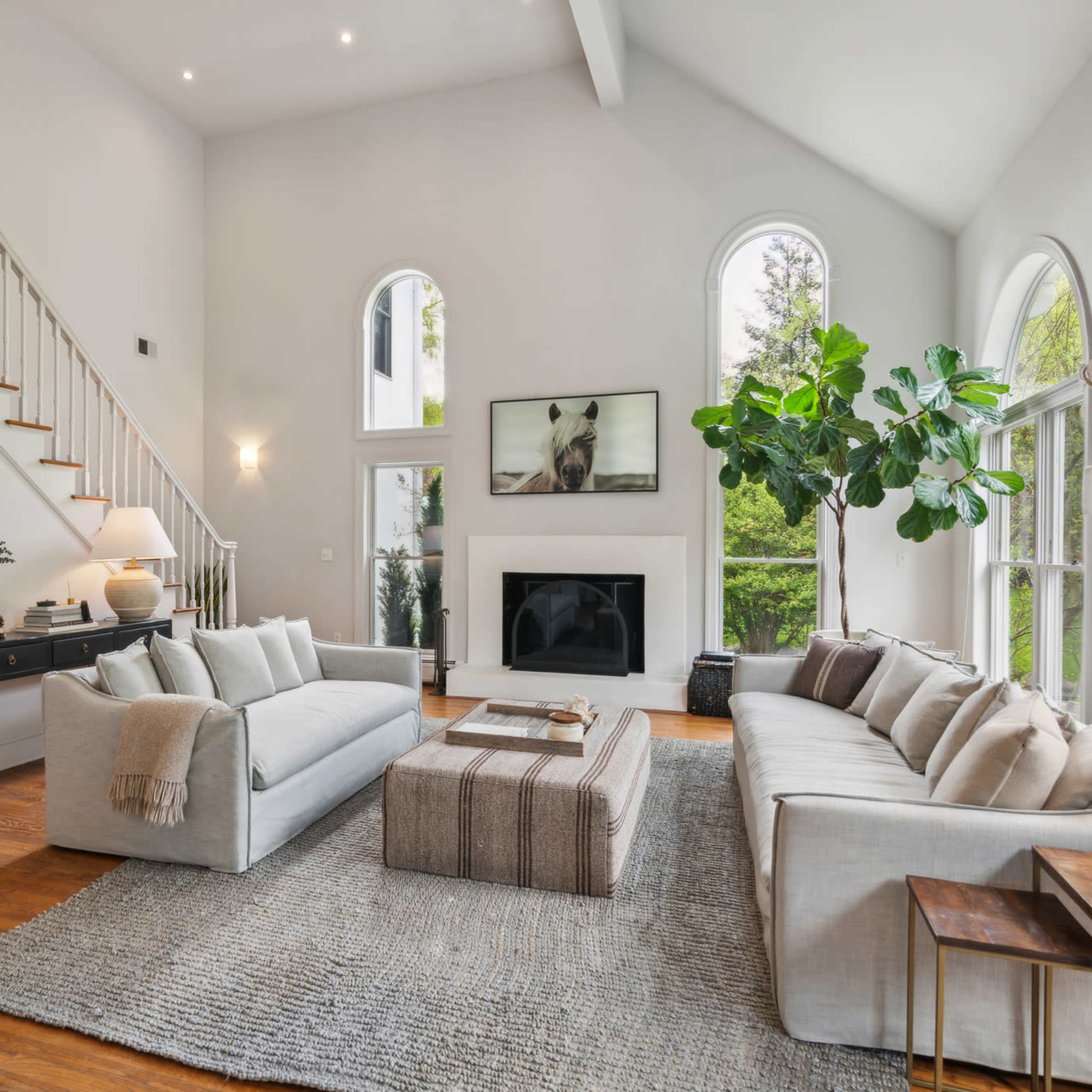 A modern living room with large windows, a beige sectional sofa, a coffee table, and a staircase leading to an upper level.