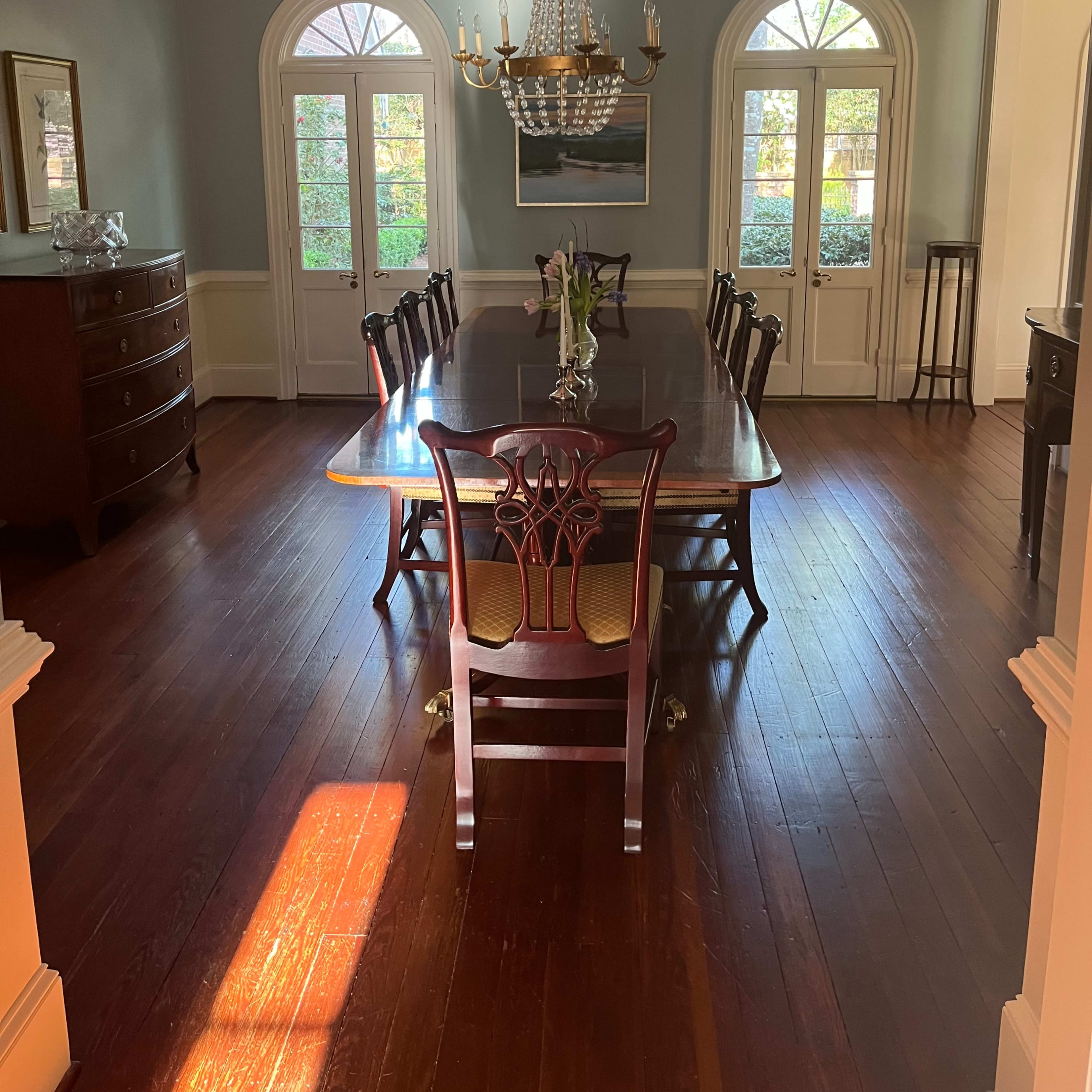 A spacious dining room with a large wooden table surrounded by chairs, a chandelier above, and large windows letting in natural light.