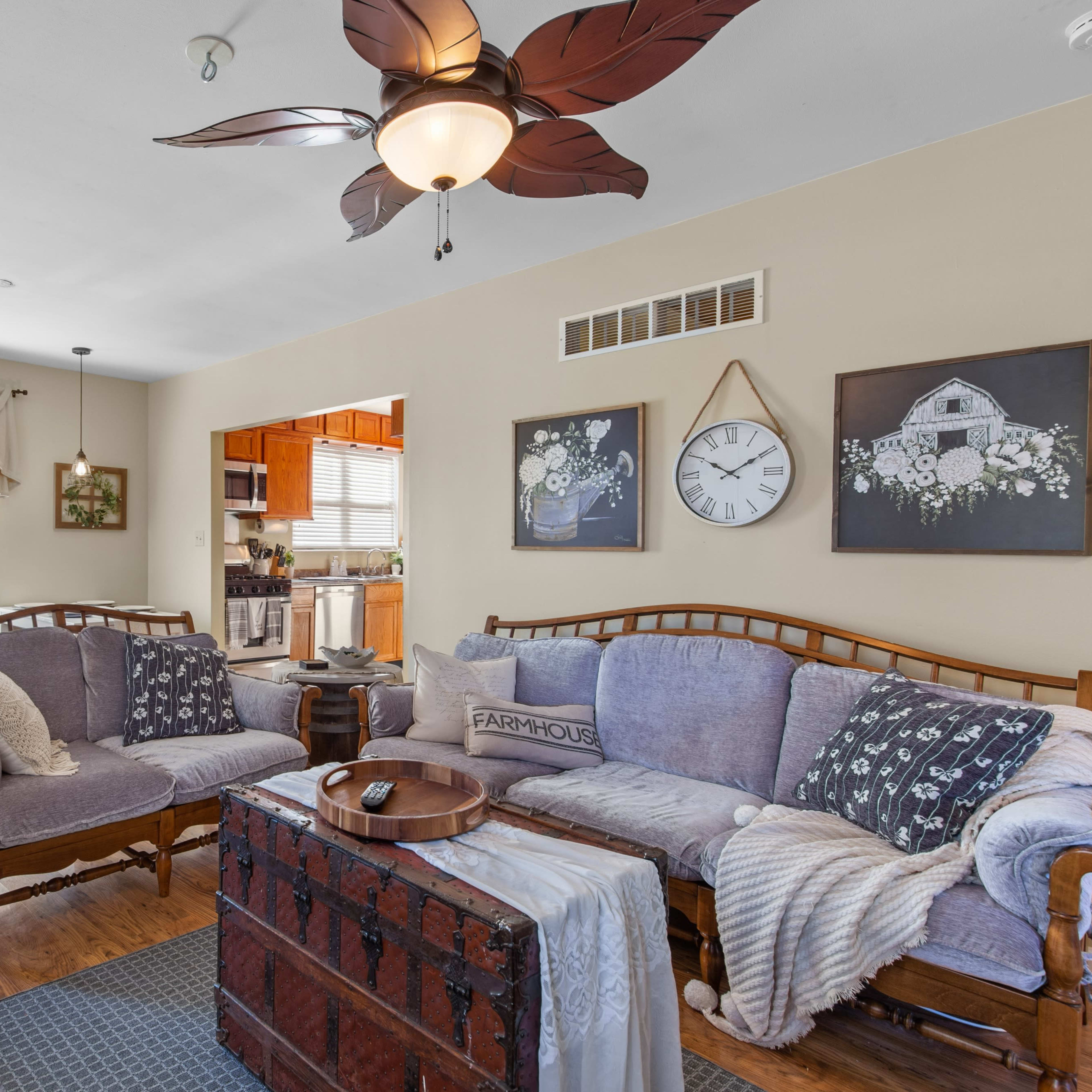 A cozy living room featuring a pair of wooden couches, a central trunk table, and decor including wall art, a clock, and a dining area visible in the background.