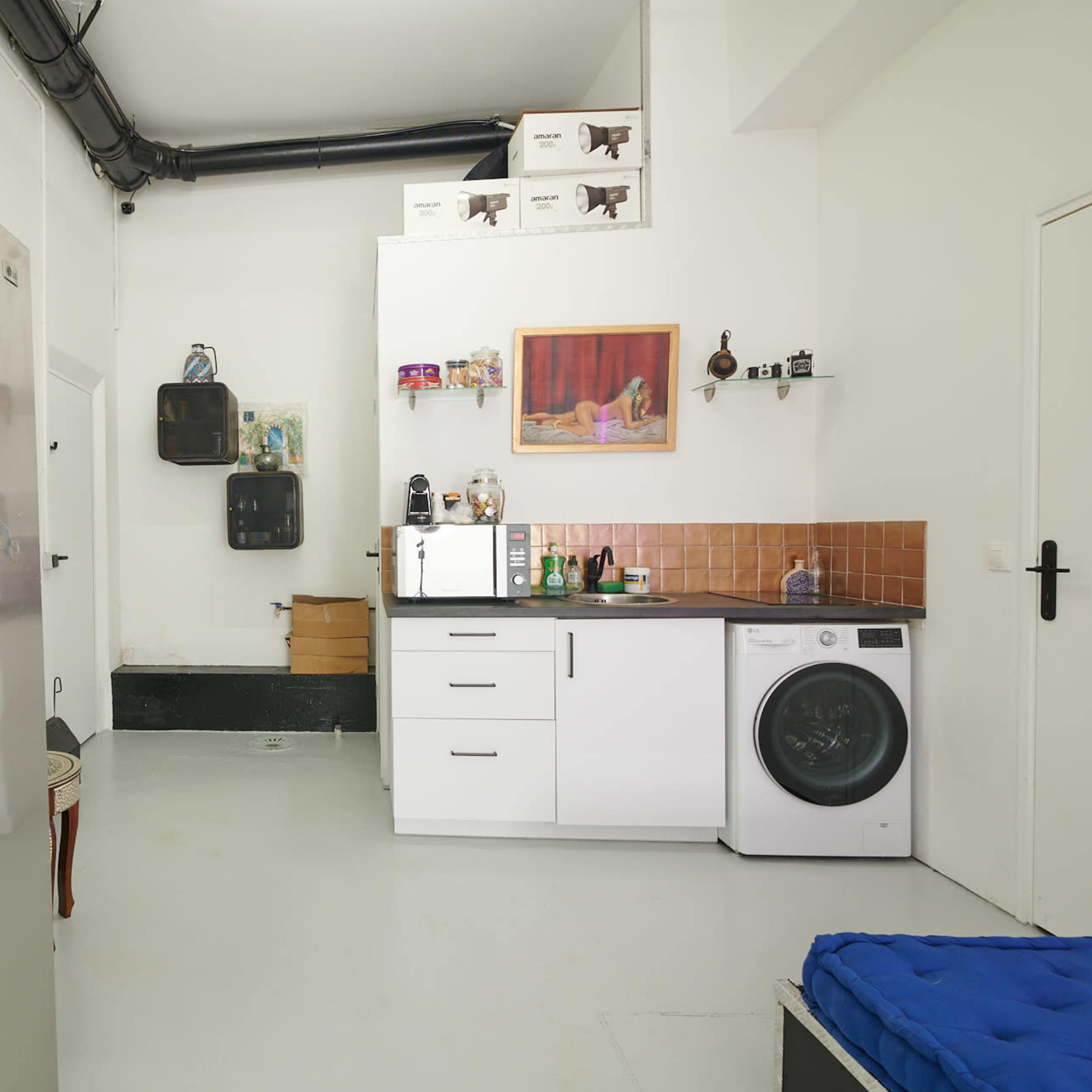 A kitchenette with a refrigerator, washing machine, and shelving is positioned in a simple room with white walls and a tiled backsplash.