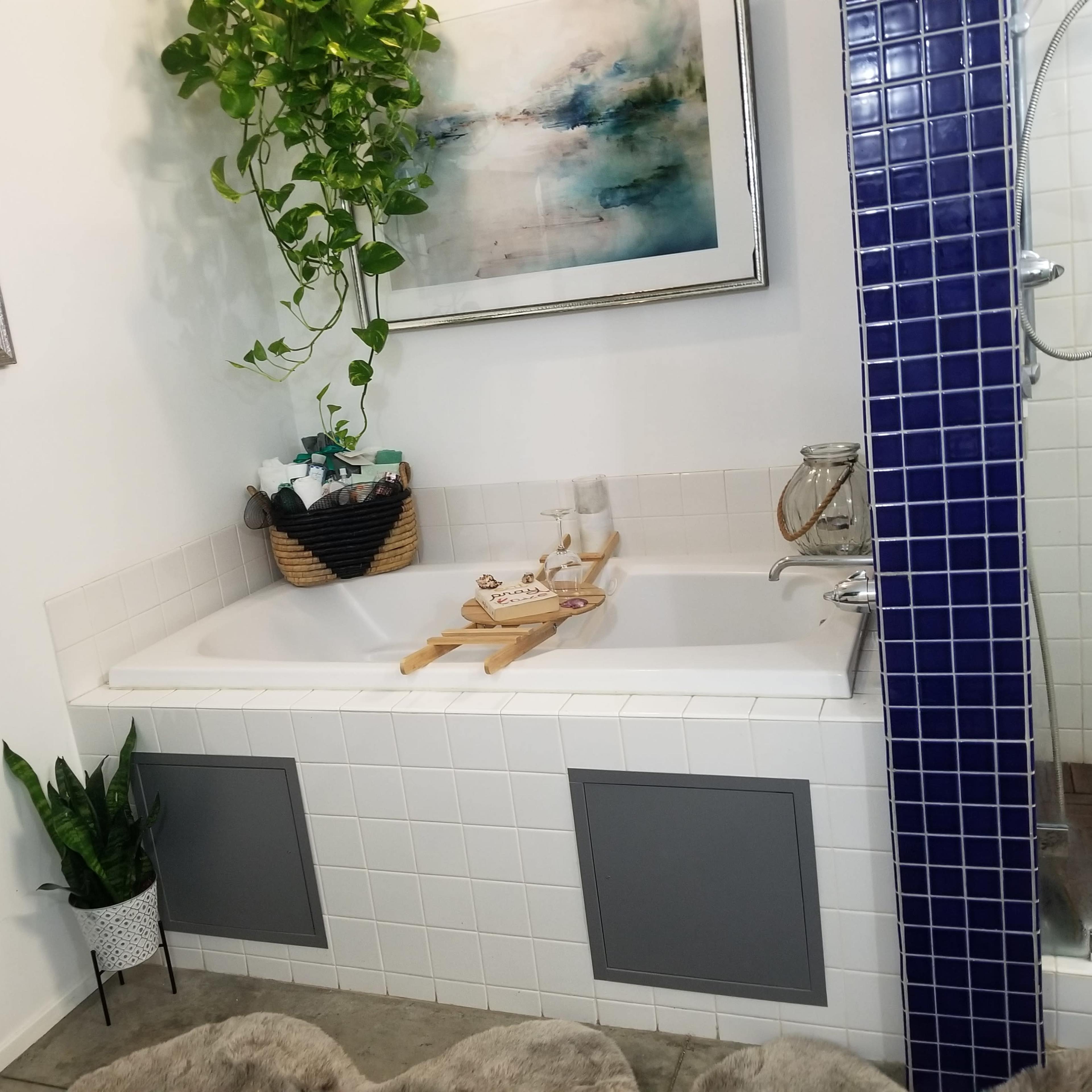 A bathroom featuring a white bathtub with a wooden tray, a shower area with blue tile, and a wall adorned with a plant and artwork.