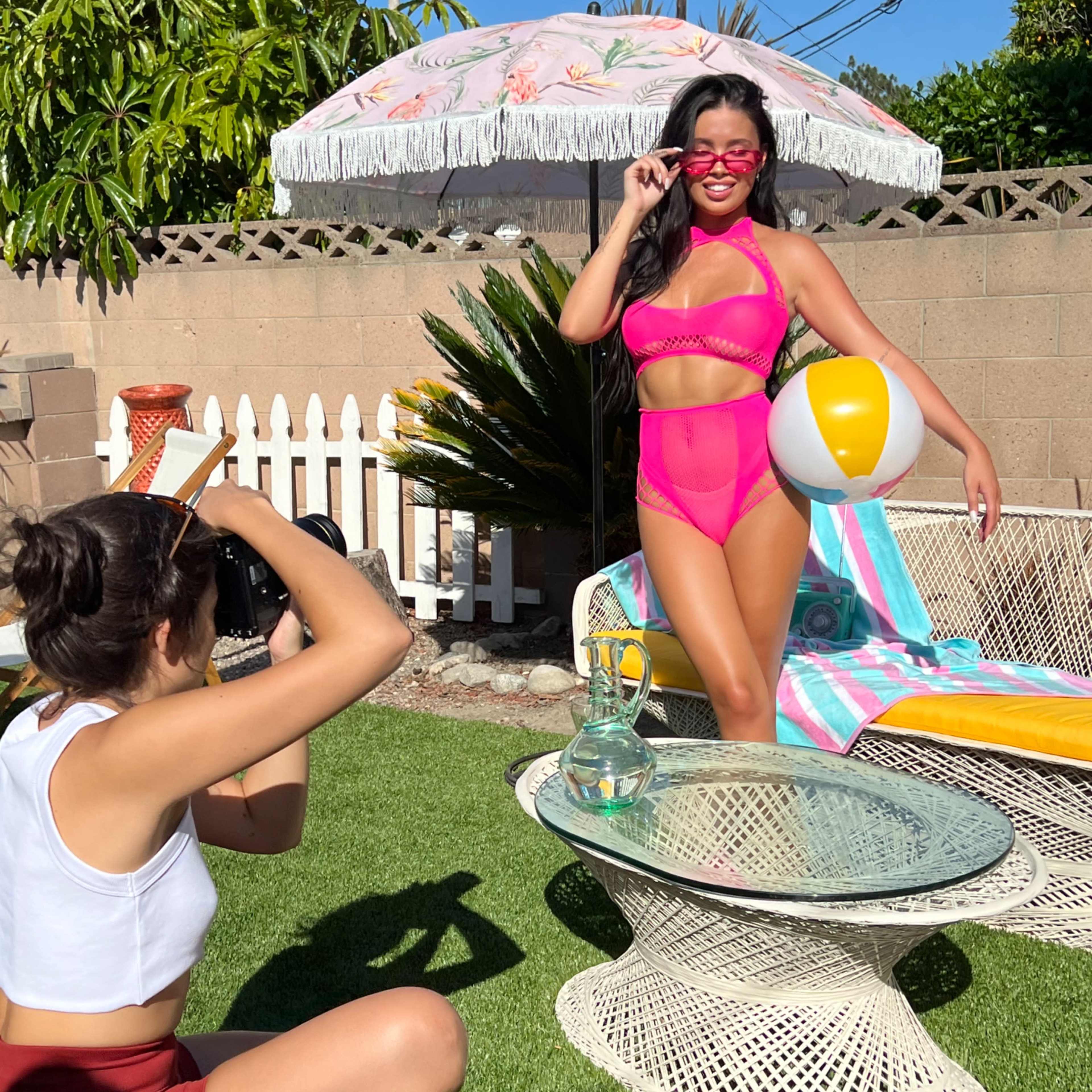 A photographer captures a model posing in a pink swimsuit by a poolside with a beach ball while a colorful umbrella provides shade.