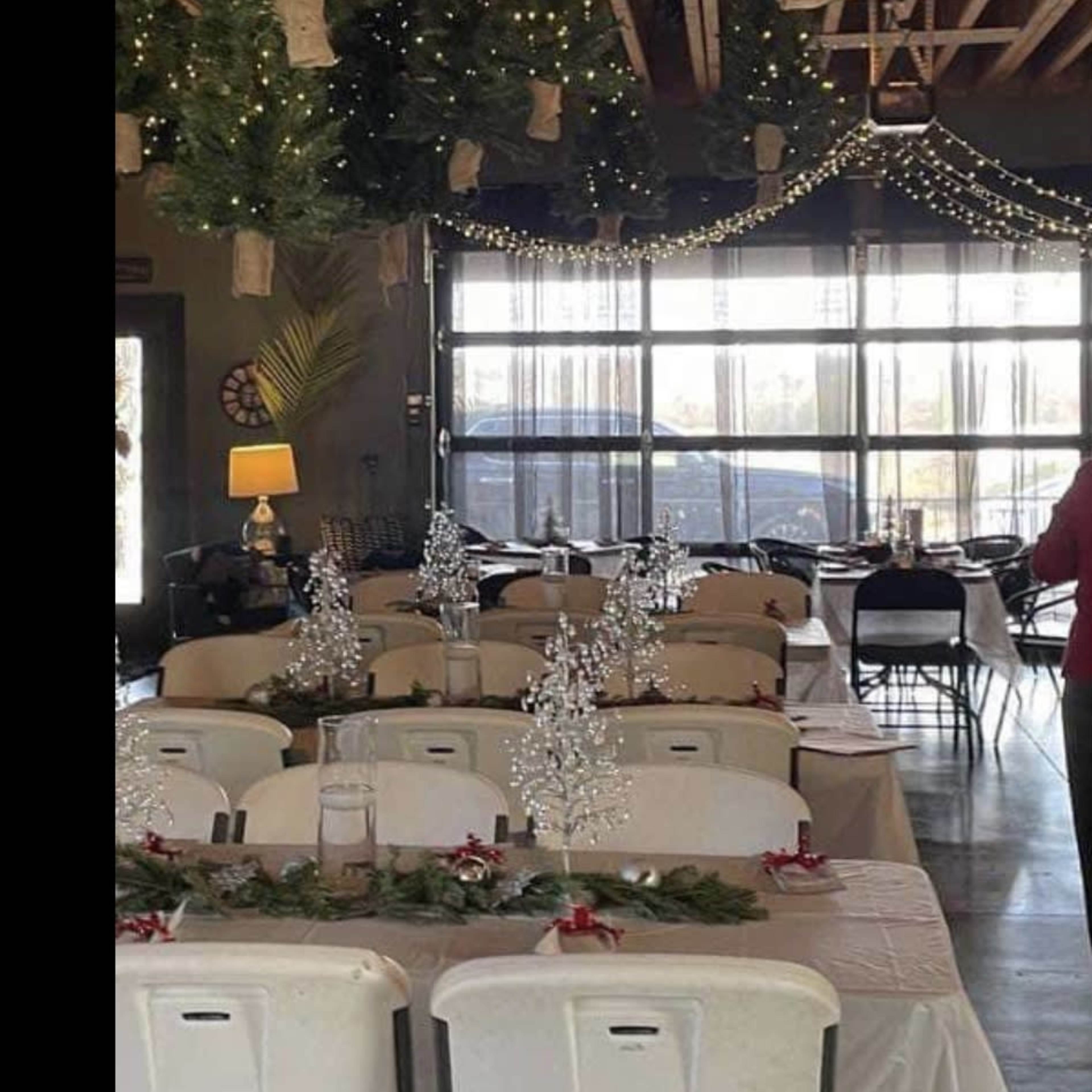 A decorated indoor dining area with tables set for a festive event, featuring hanging greenery and string lights overhead.