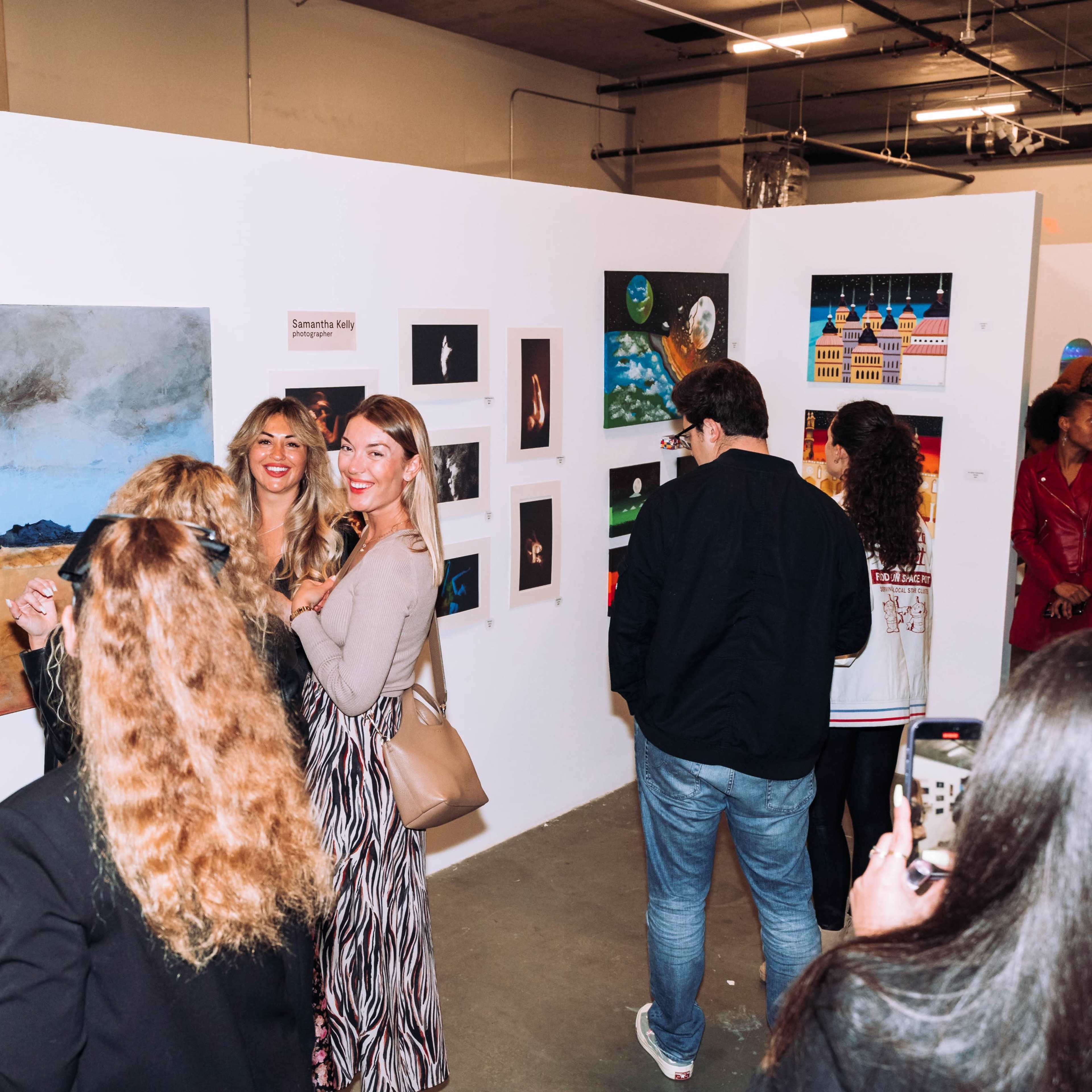 A group of people gather around an art exhibition showcasing various paintings on white walls.