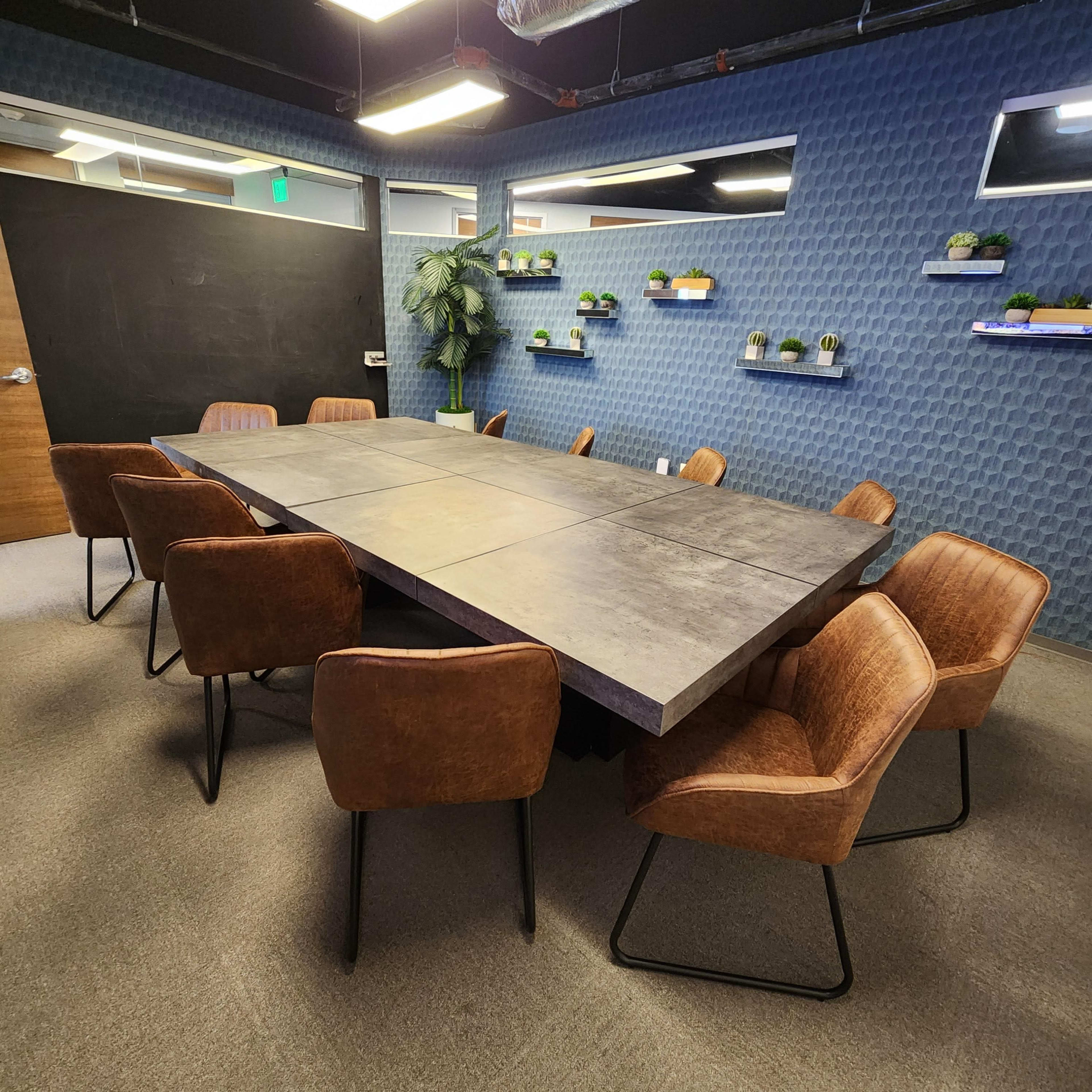 A large, rectangular conference table surrounded by eight brown chairs is positioned in a modern meeting room with blue textured walls and small potted plants on shelves.