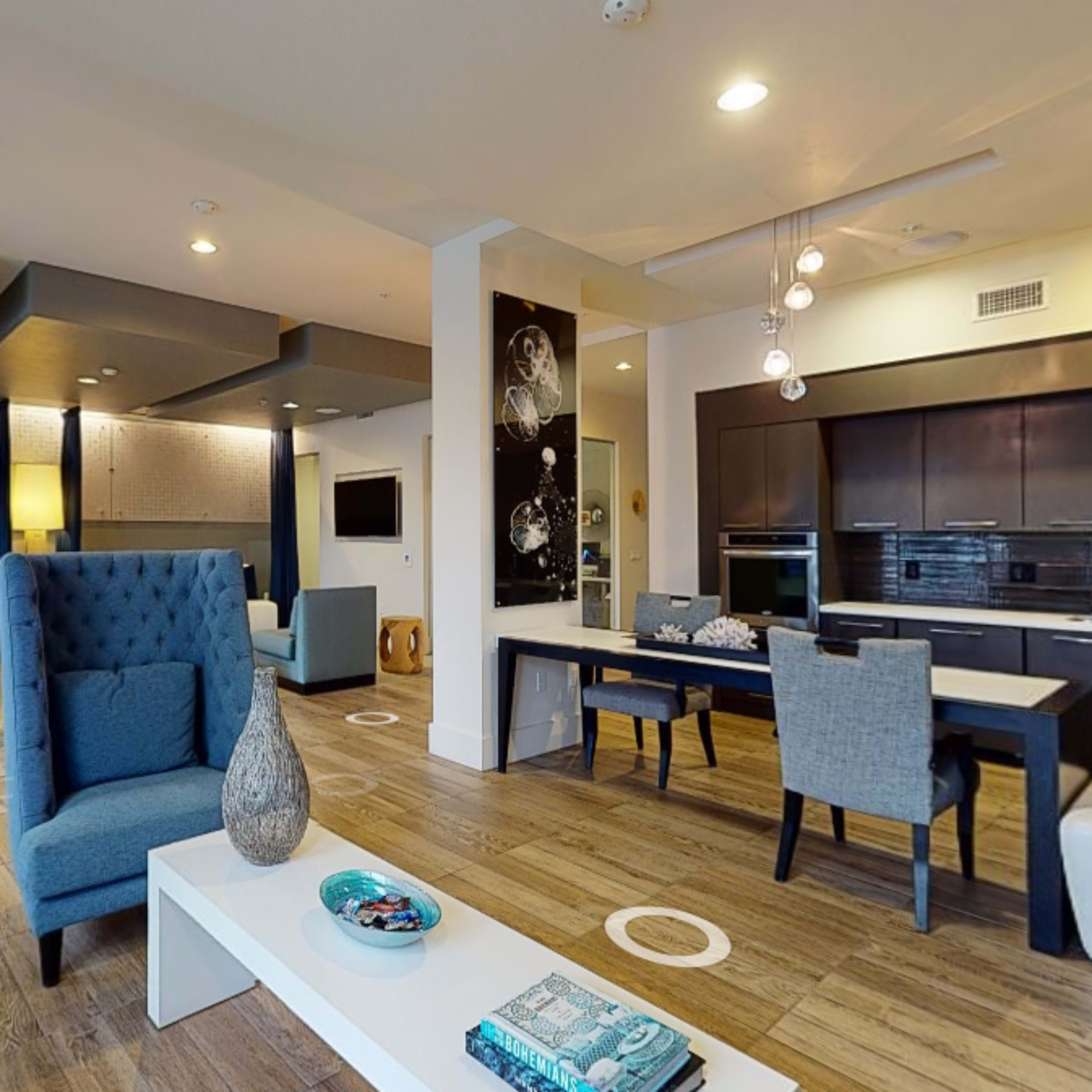 A modern living space featuring a blue armchair, a dining area with gray chairs, and a kitchen in the background, all with wooden flooring.