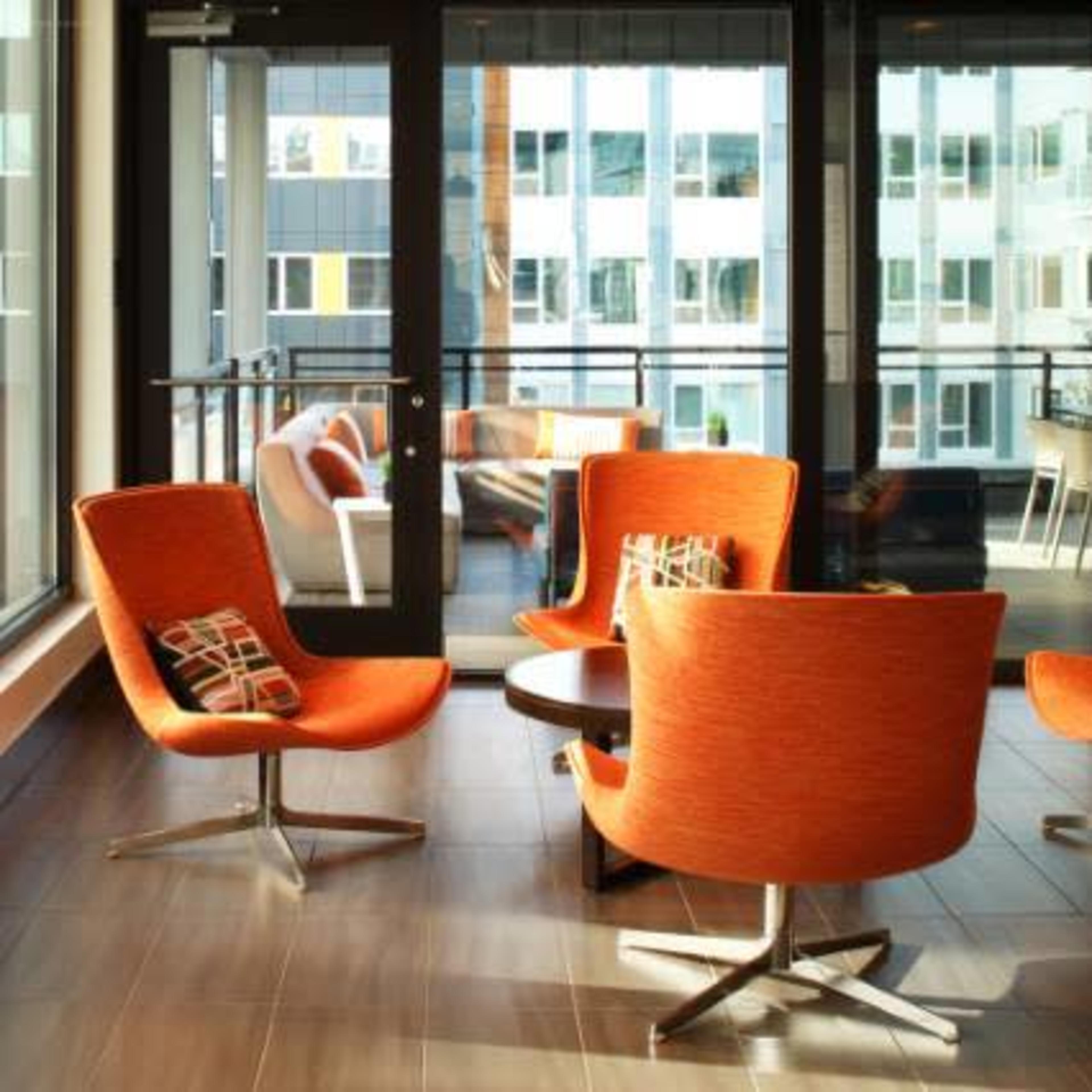 A modern lounge area with four orange chairs arranged around a circular table, surrounded by large glass windows.