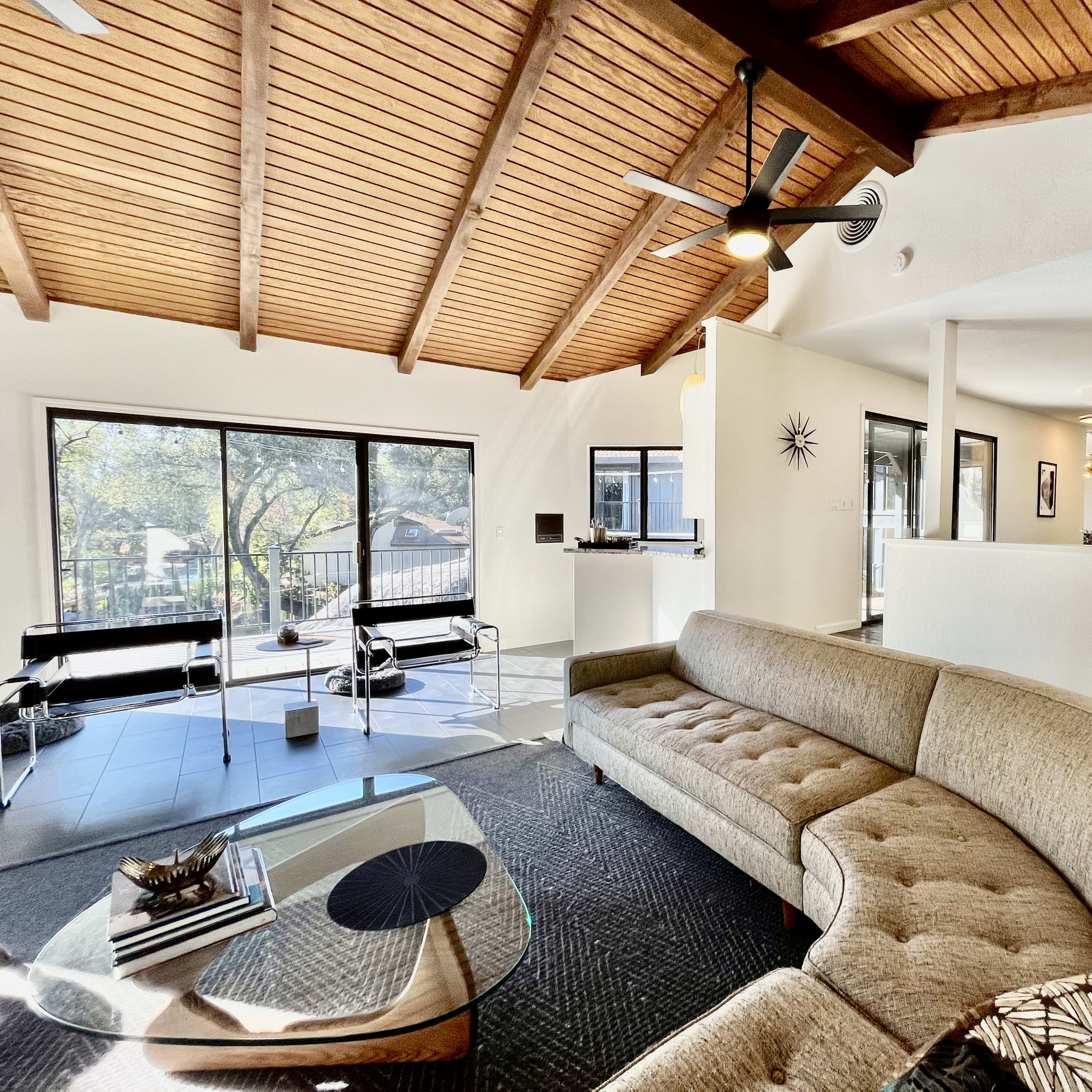 A modern living room features a curved sectional sofa, a glass coffee table, and large windows that provide natural light, all set under a vaulted wooden ceiling.