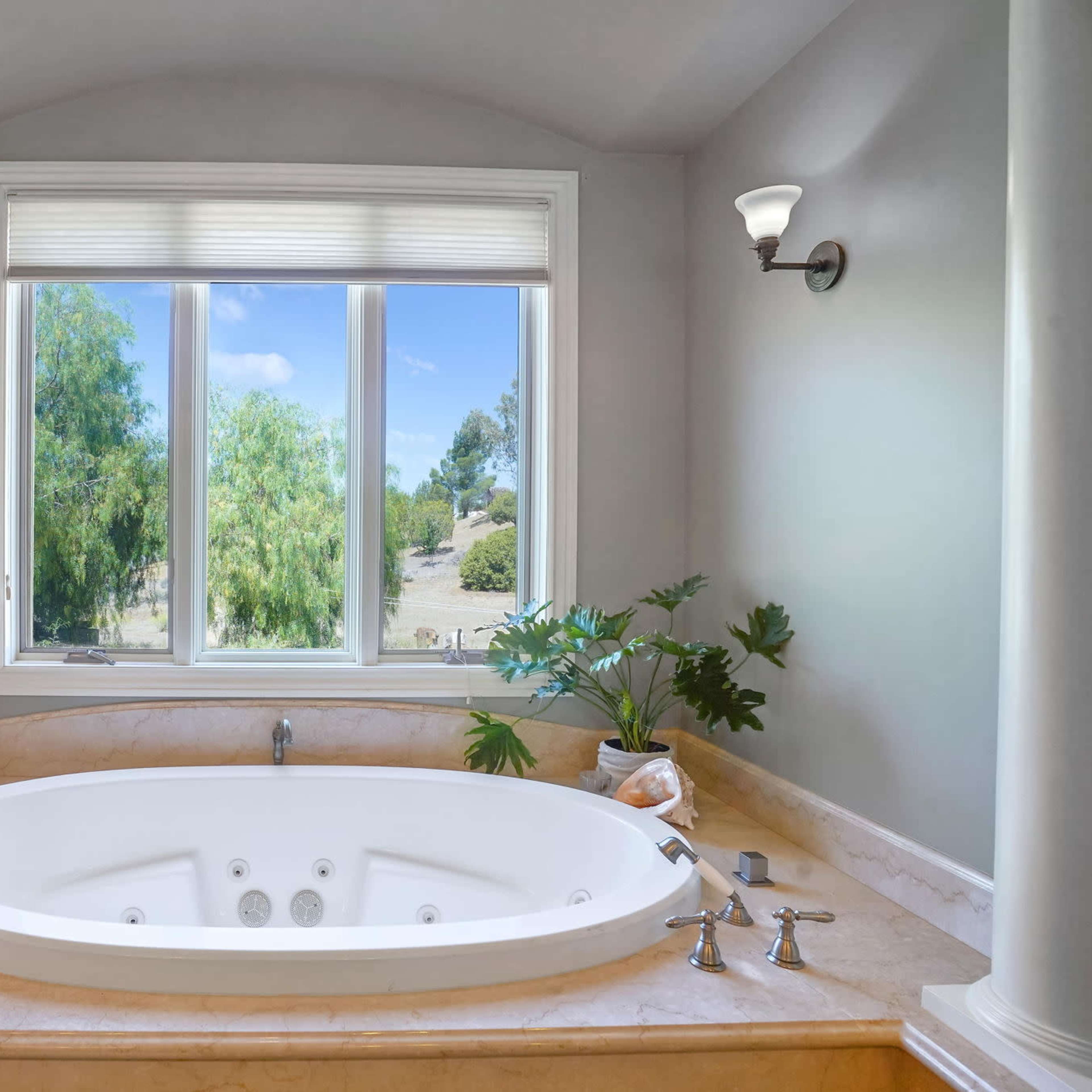 A modern bathroom features a large oval bathtub positioned near a window with a view of greenery outside.