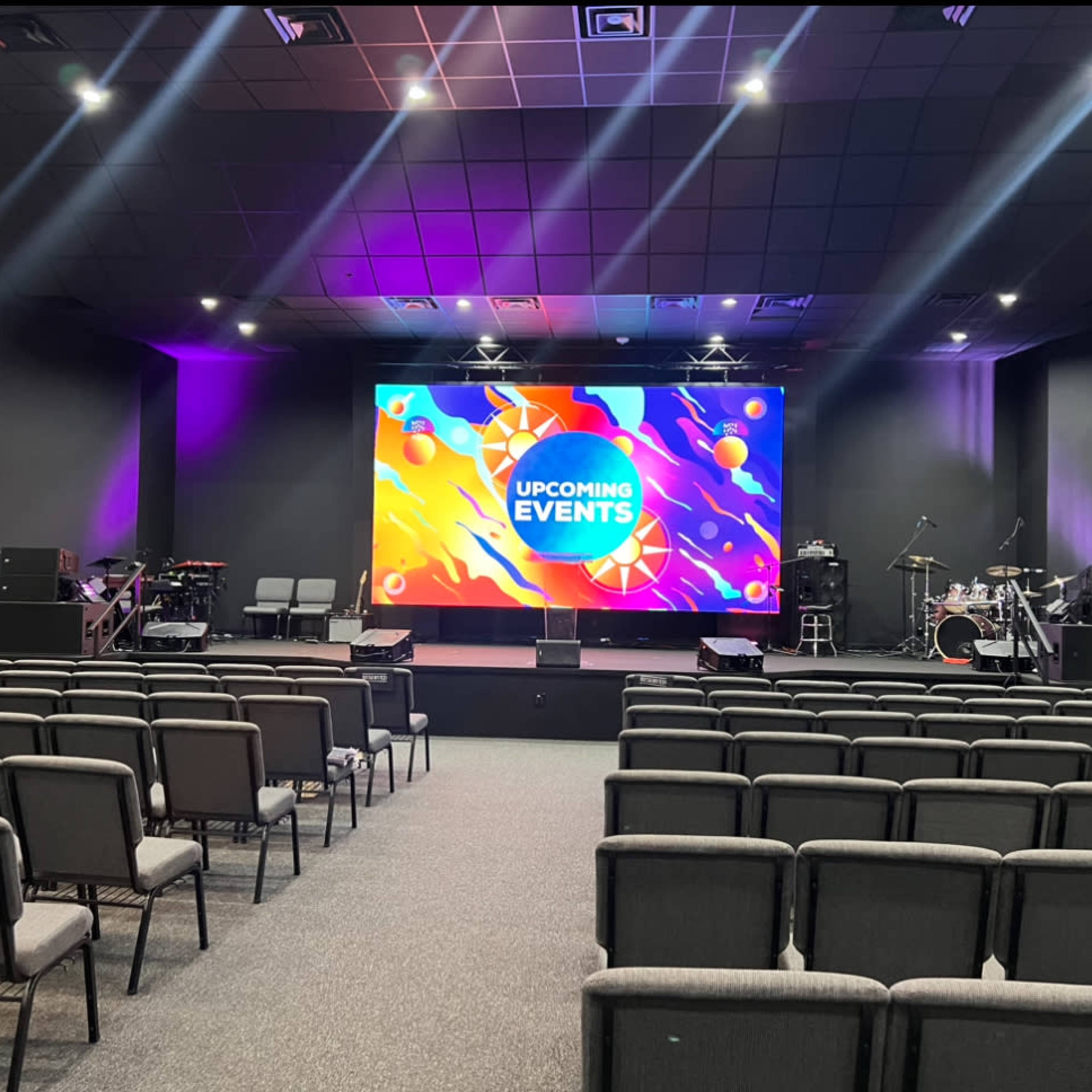 A performance space with several rows of gray chairs facing a large screen displaying "UPCOMING EVENTS," alongside musical instruments set up on stage.