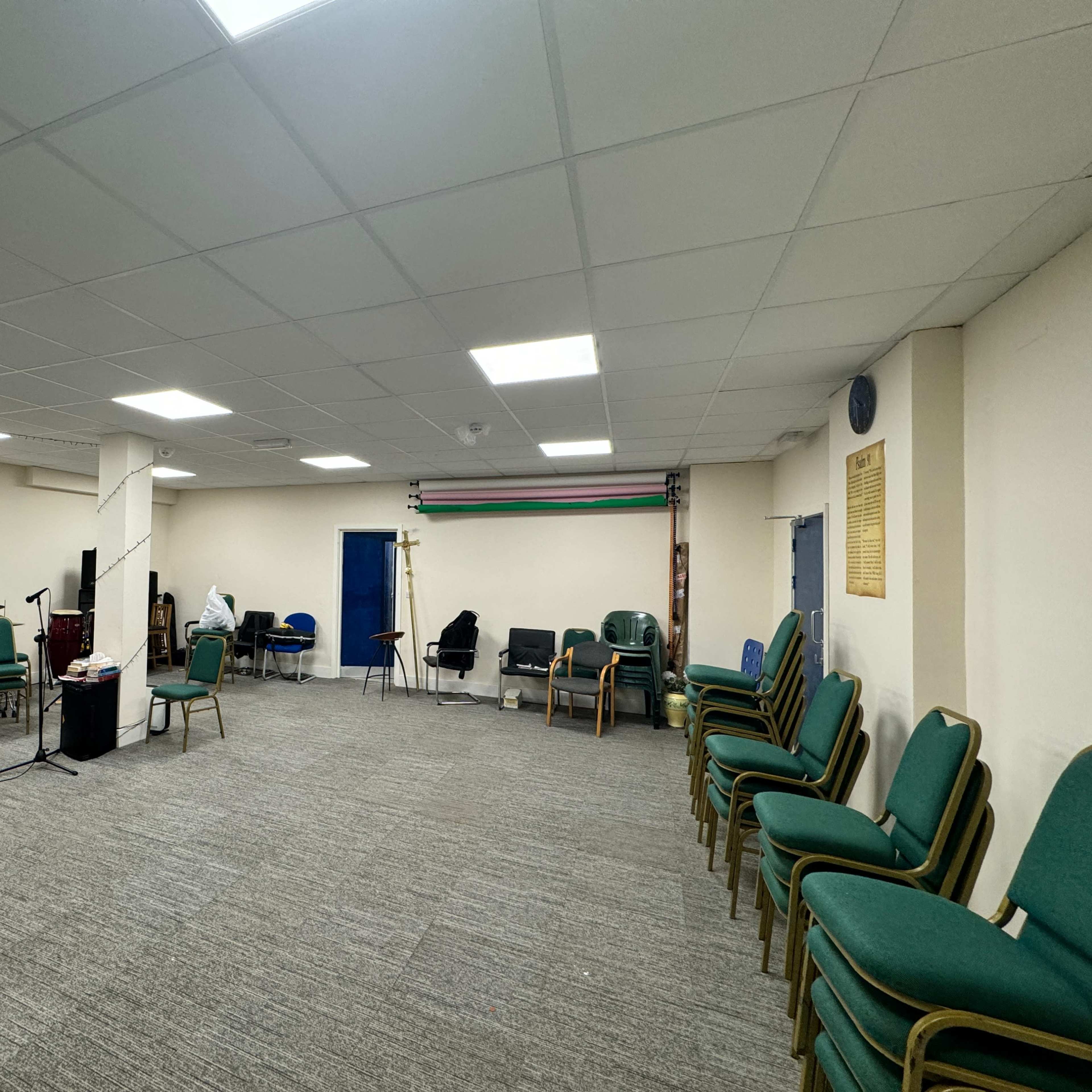 A spacious room equipped for music practice, featuring a drum set, several chairs, and a blue door leading to another area.