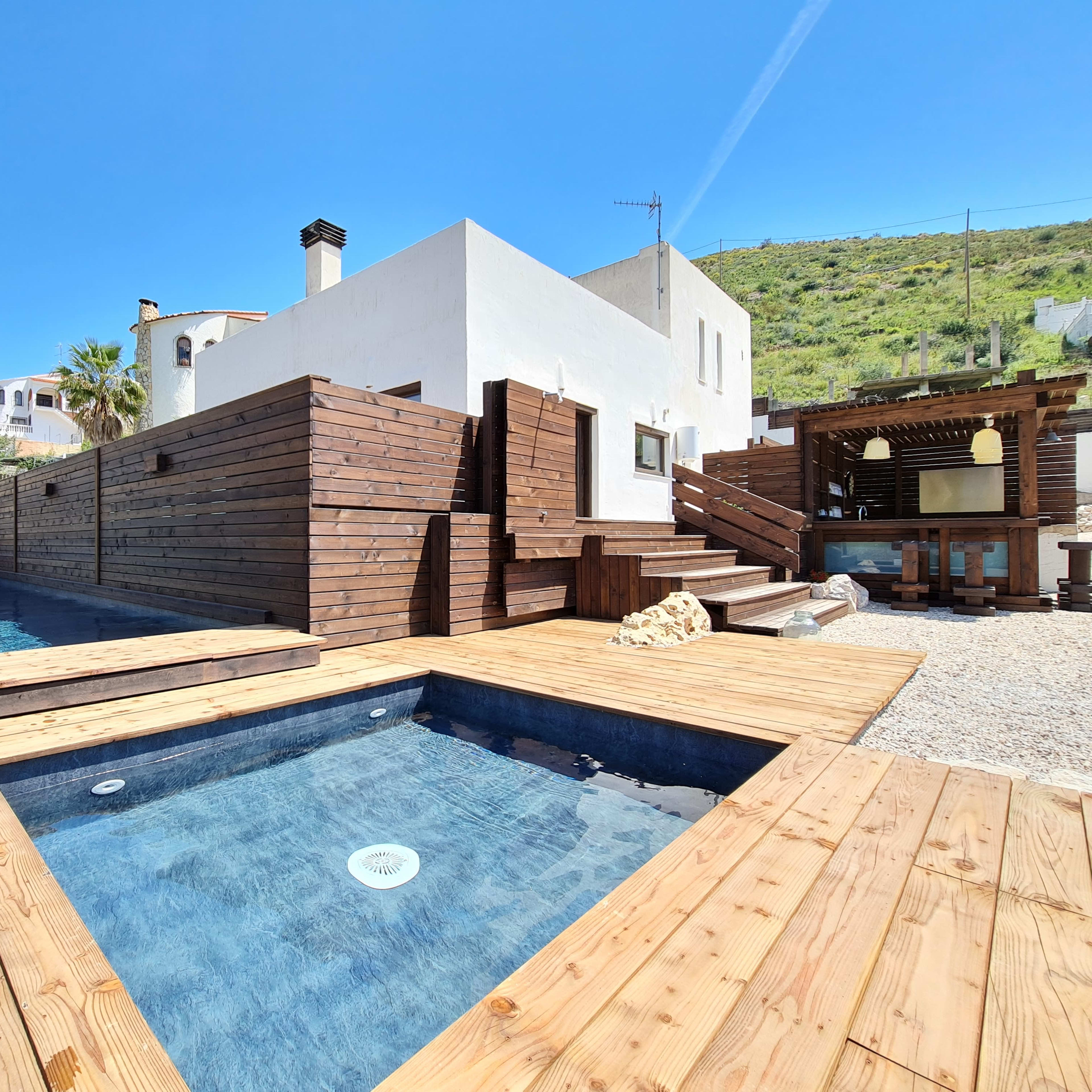 A modern house with wooden decking, featuring two small pools surrounded by a landscaped area under a clear blue sky.