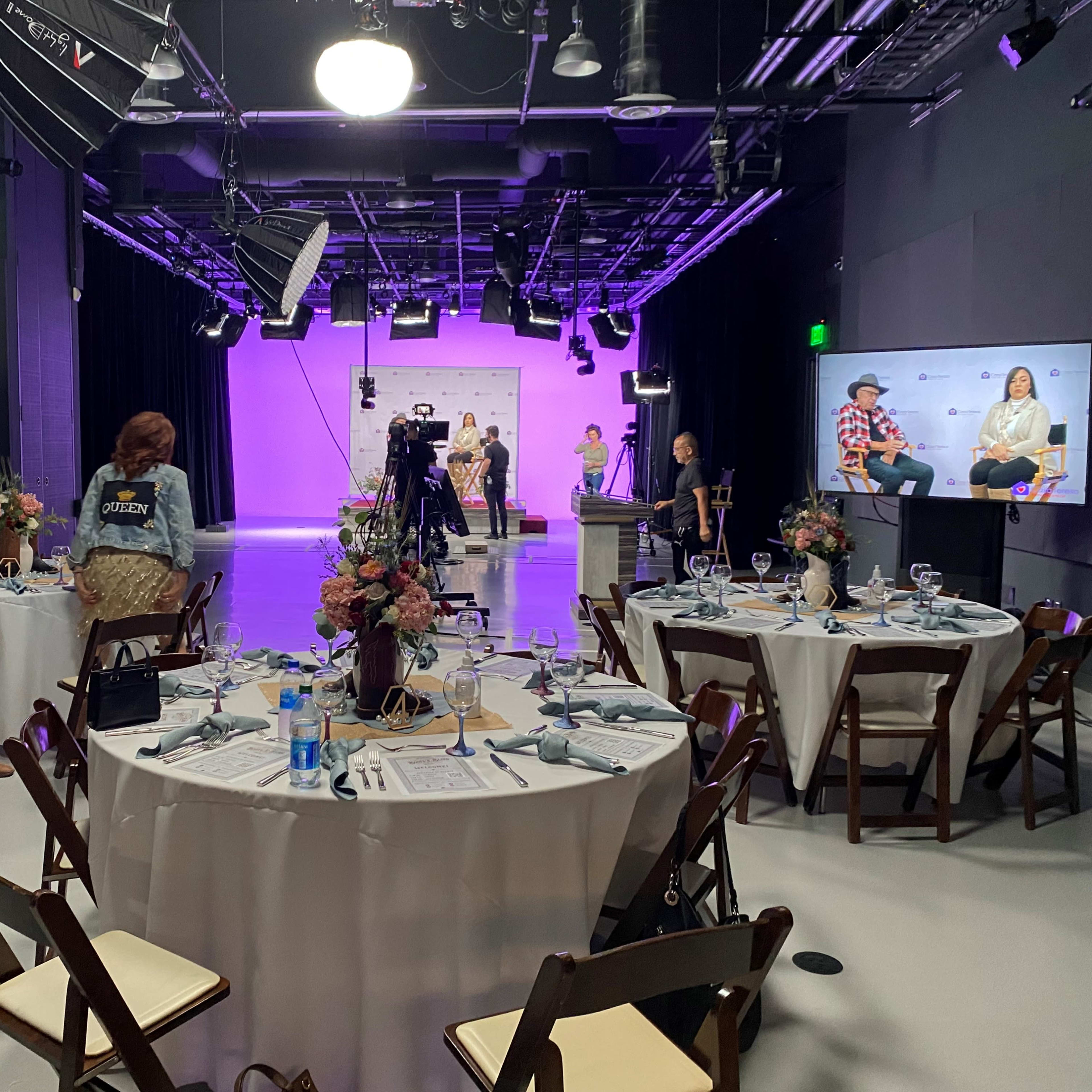 A television studio is set up for an event, featuring tables arranged for dining and a stage with speakers and a camera crew.