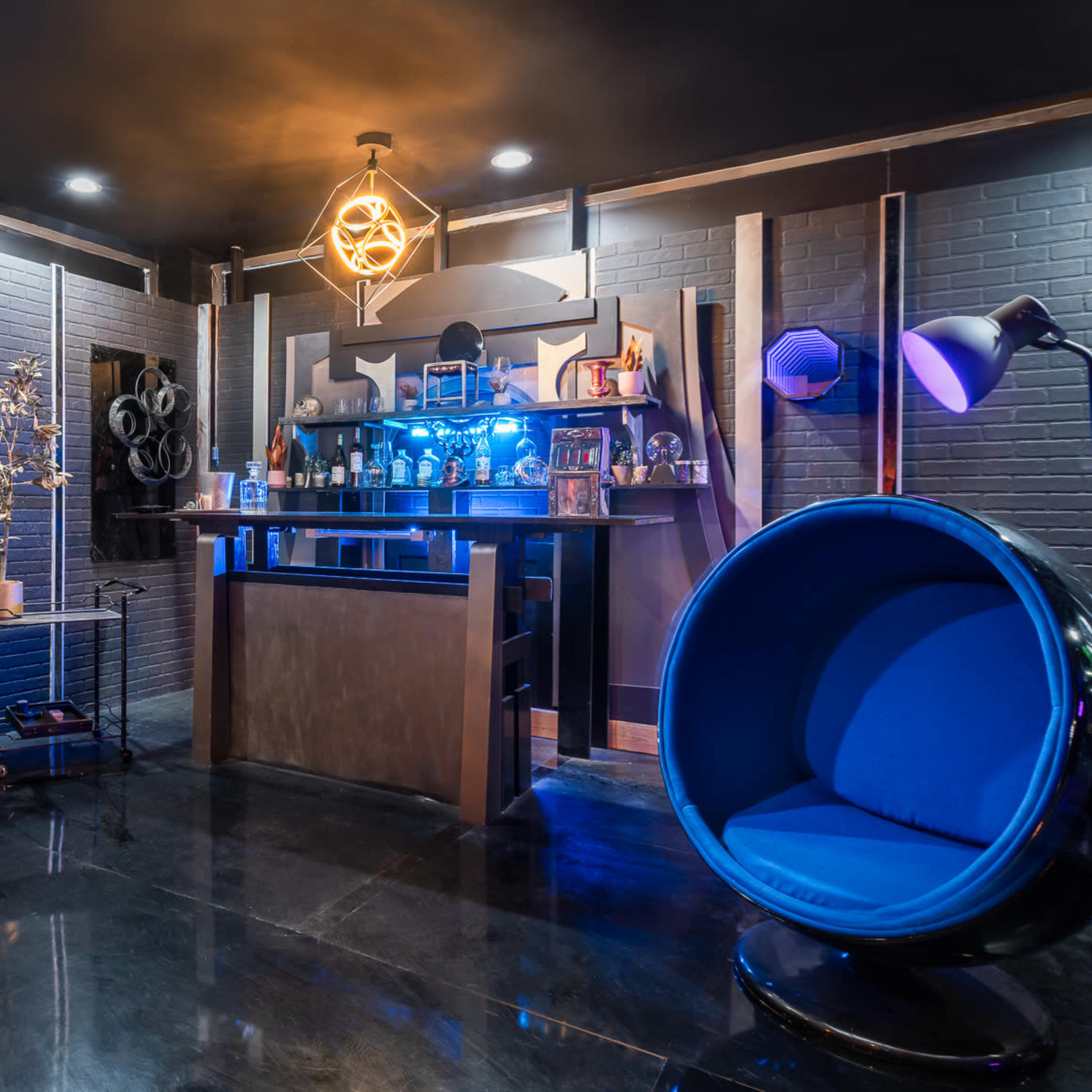 A stylish bar area with a round blue chair, illuminated shelves filled with decorative items, and a metallic structure resembling a door or entryway.