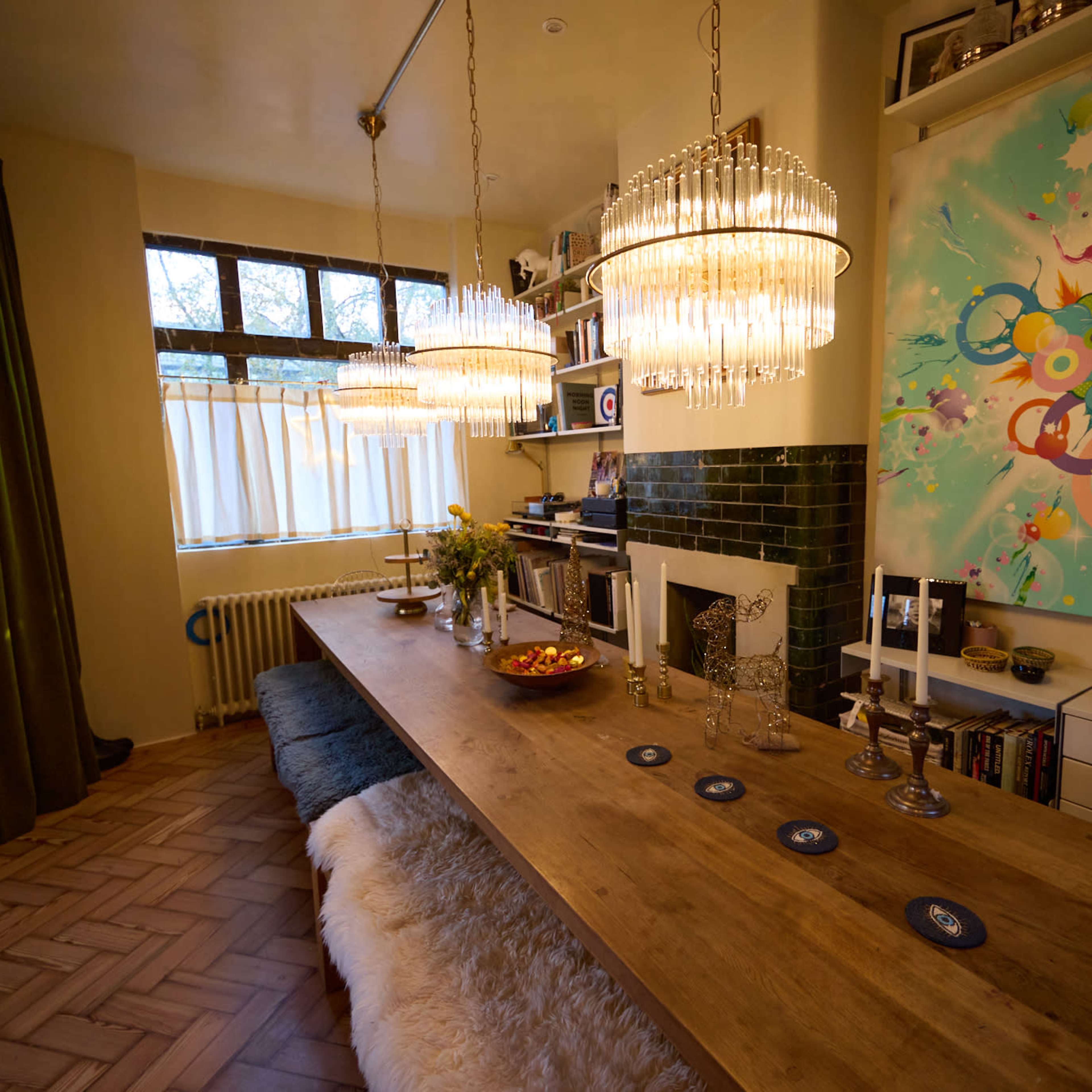 A spacious dining room features a wooden table with crystal pendant lights, a decorative wall with colorful artwork, and a green tiled fireplace.