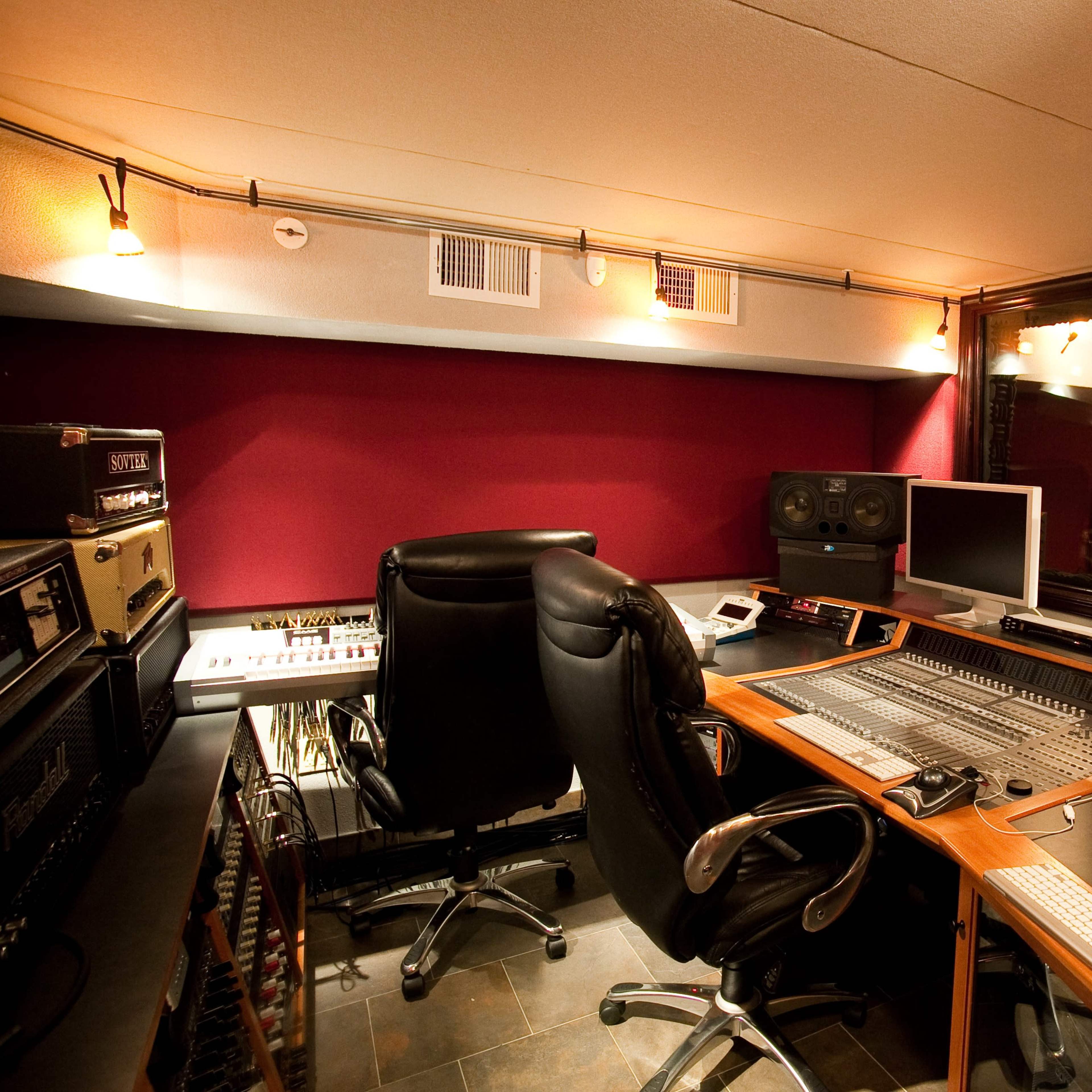 A music recording studio with a control console, computers, sound monitors, and various audio equipment arranged around the room.