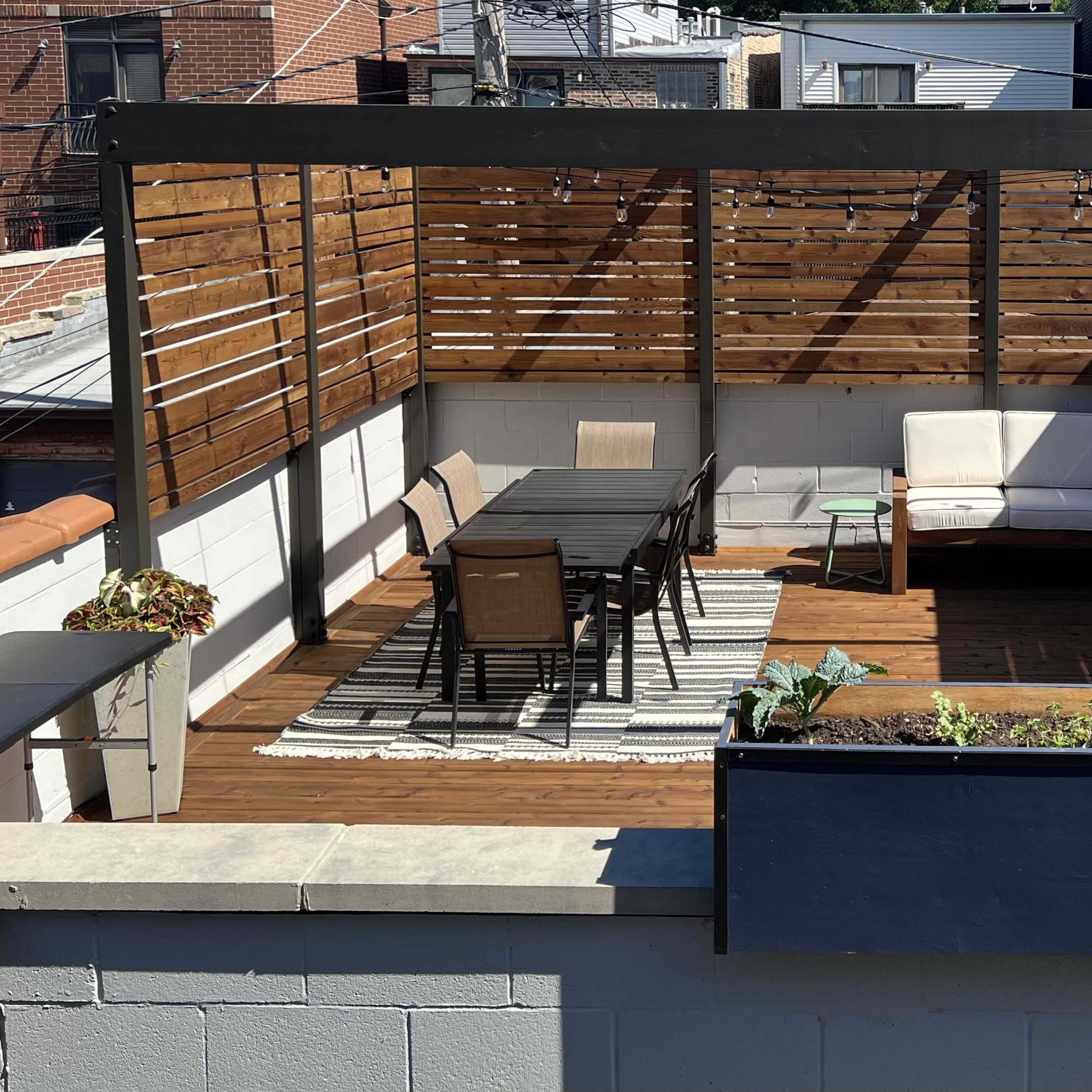 A rooftop terrace featuring a wooden pergola, outdoor seating, a dining table, and potted plants.